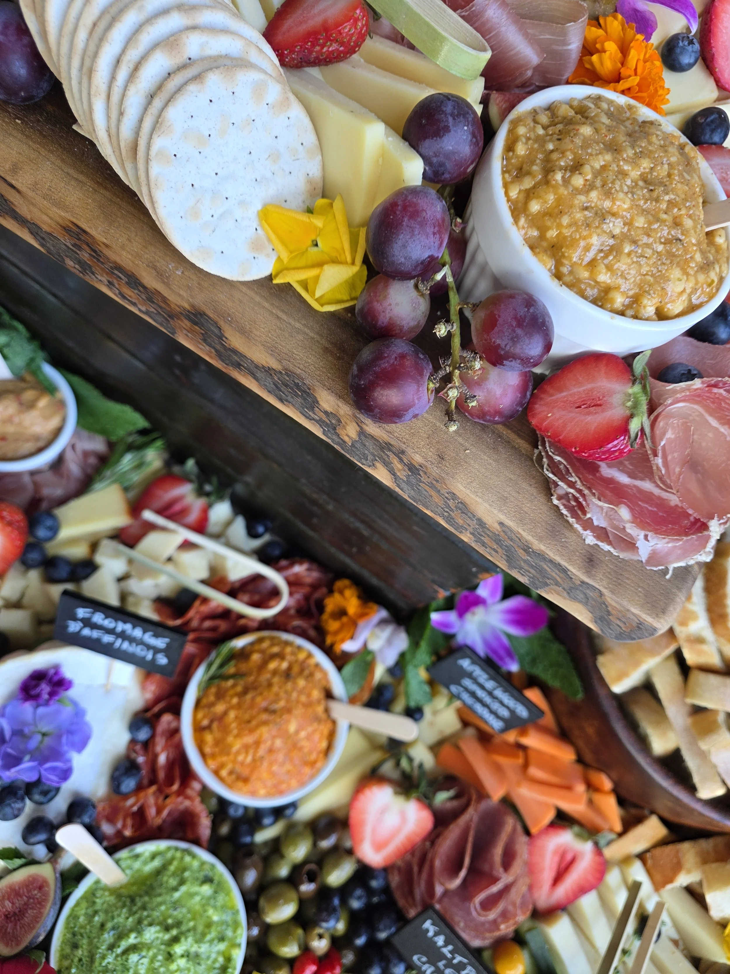A charcuterie board with sliced cheese, grapes, strawberries, blueberries, figs, cured meats, and a bowl of grain mustard, decorated with edible flowers.