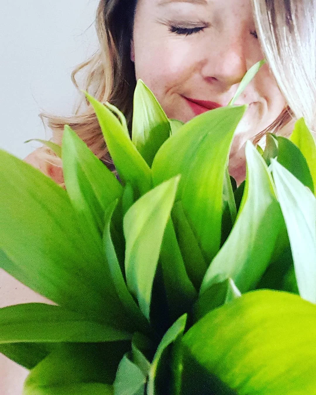 A woman with blonde hair and closed eyes holding a large green bouquet of fresh forged wild leeks or ramps. Enjoy the simplicity of local naturally available ingredients.