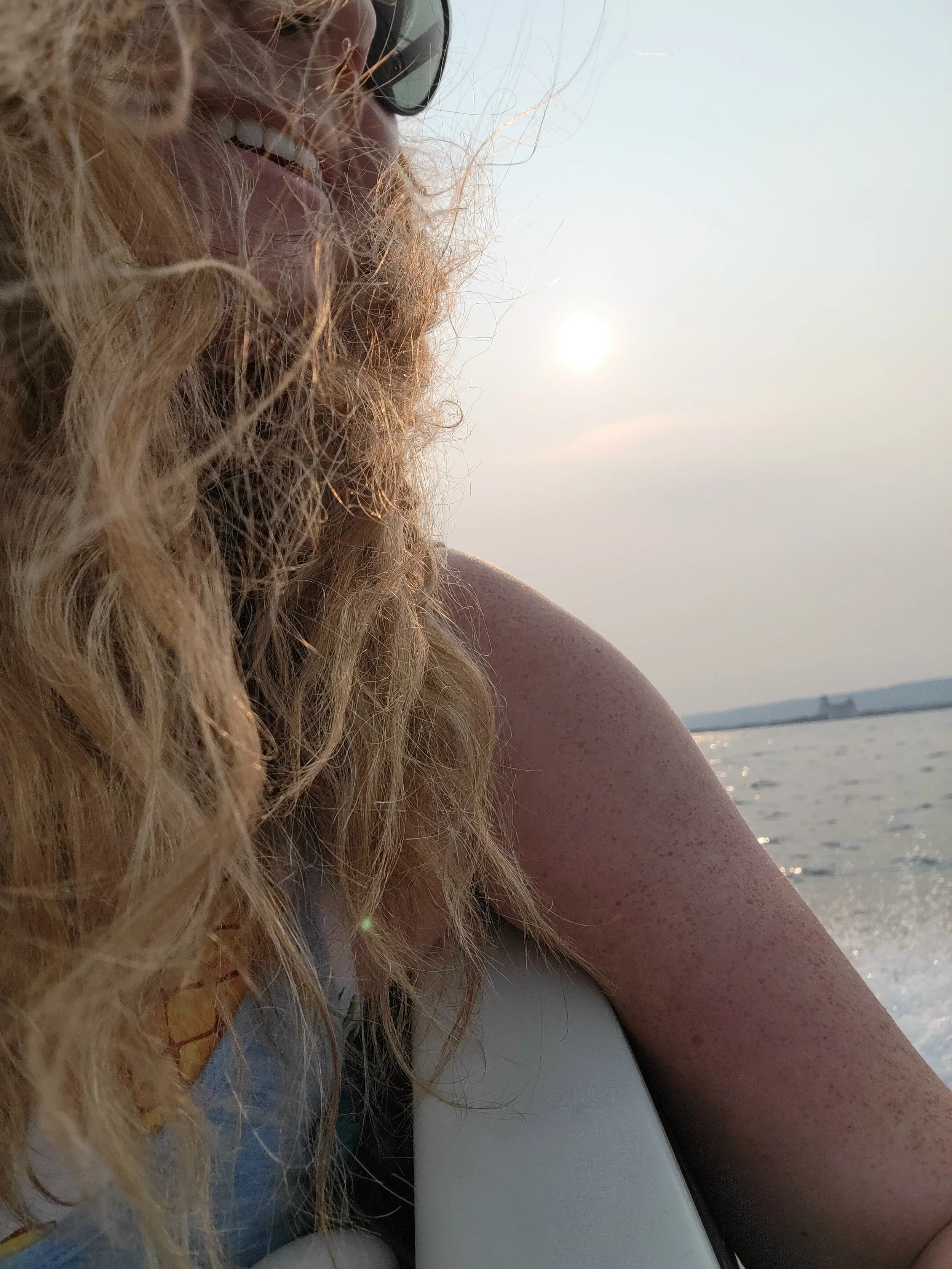 Person with curly blonde hair, wearing sunglasses, smiling, near water at sunset or sunrise. Lakeside fun, life on the bay, enjoy the water, smiles, georgian bay