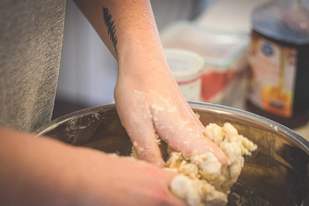 Person mixing dough in a metal bowl with their hands during baking, with containers of ingredients in the background.