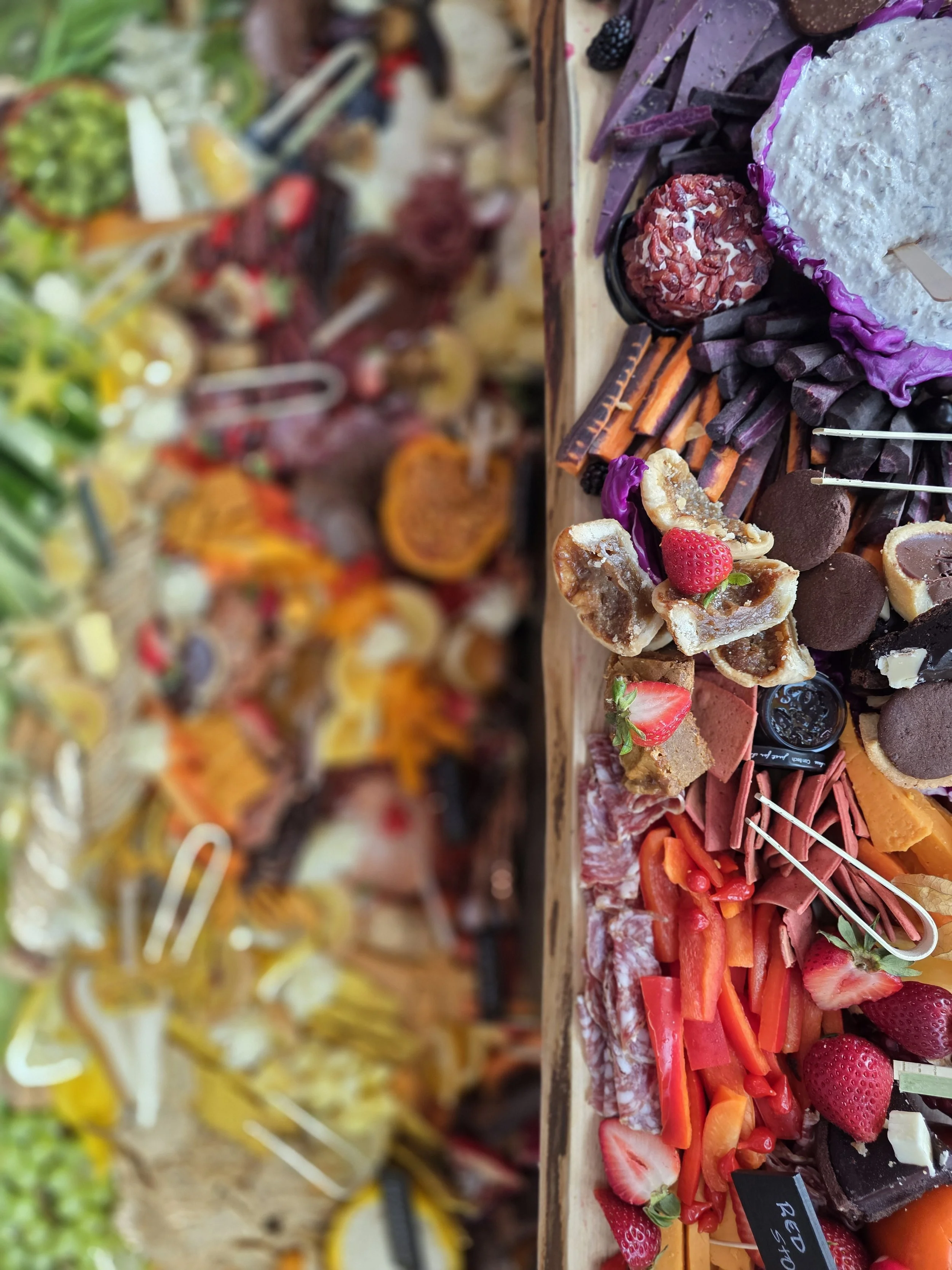 A variety of desserts and snacks on display, including strawberries, chocolate treats, sliced meats, and colorful confections, arranged on a long table at a market or celebration.