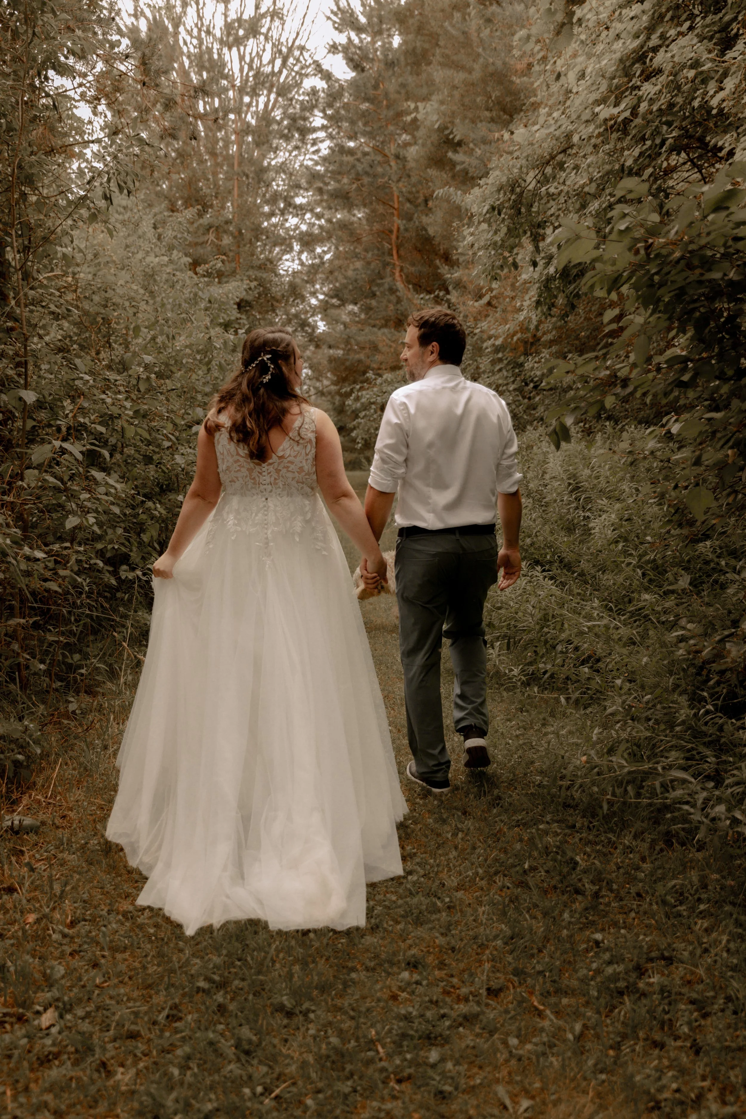 A newlywed couple walking hand in hand through a wooded trail, dressed in wedding attire with the bride in a wedding dress and the groom in a white shirt and dark pants, surrounded by lush greenery and tall trees. family, togetherness, love, wedding.
