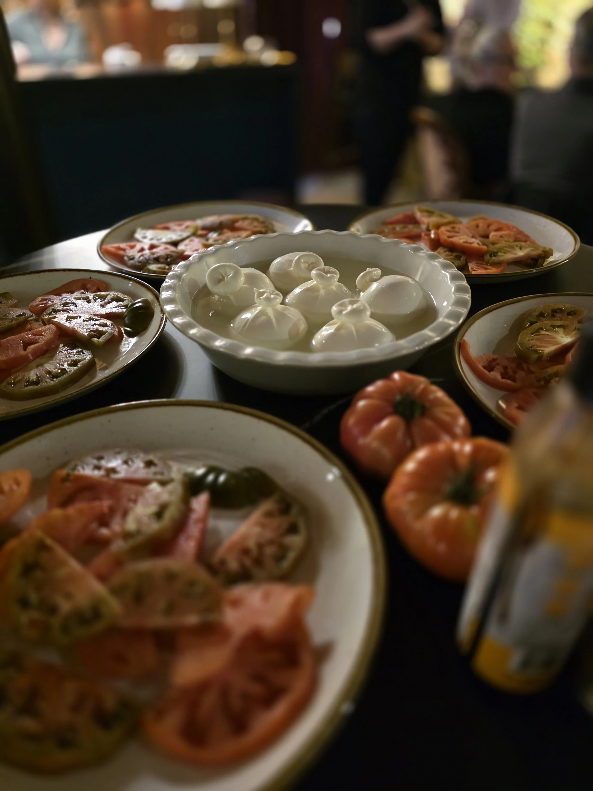 Plates of sliced fresh vine and heirloom tomatoes, burrata cheese, and other vegetables on a table with blurred background. Part of a wine and food pairing showcasing Italian cheese and summer entertaining.