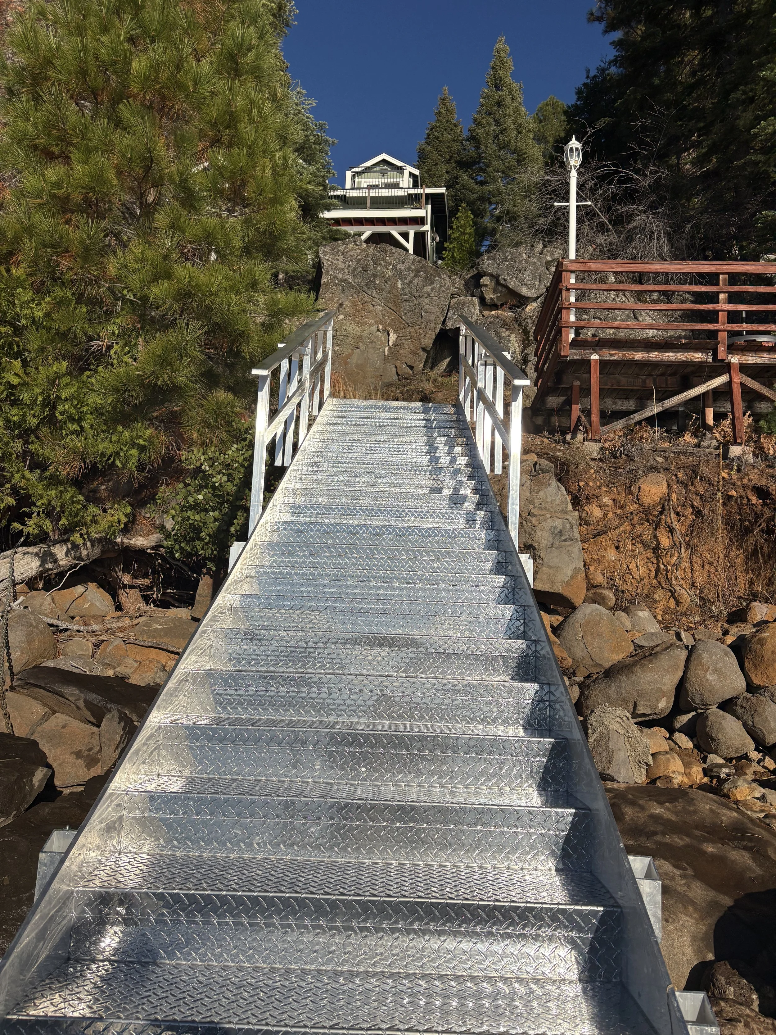 A metal footbridge with railings crossing over rocks and dry trees, leading to a house or cabin on stilts surrounded by pine trees against a clear blue sky.