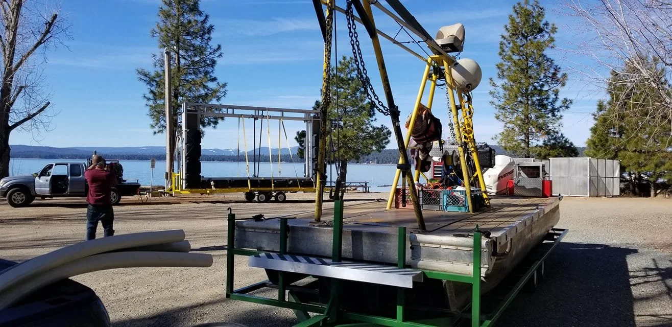 Outdoor scene with a floating platform, a baseball swing set structure, and a small stage under construction near a body of water, with trees and a clear blue sky in the background.