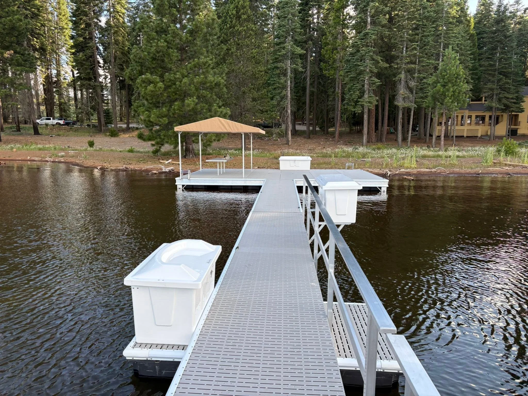 A floating dock with a canopy and storage boxes extends into a lake, with dense tall trees and a yellow house visible on the shoreline in the background.