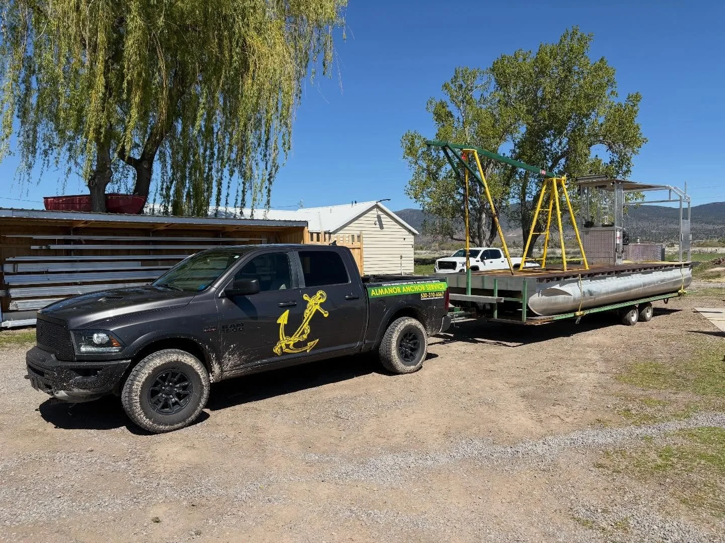 A black pickup truck with a yellow anchor logo on the side is attached to a large flatbed trailer. The trailer has a metal frame structure and a swing set and small boat on it, parked outdoors on a gravel surface under trees with a clear blue sky.