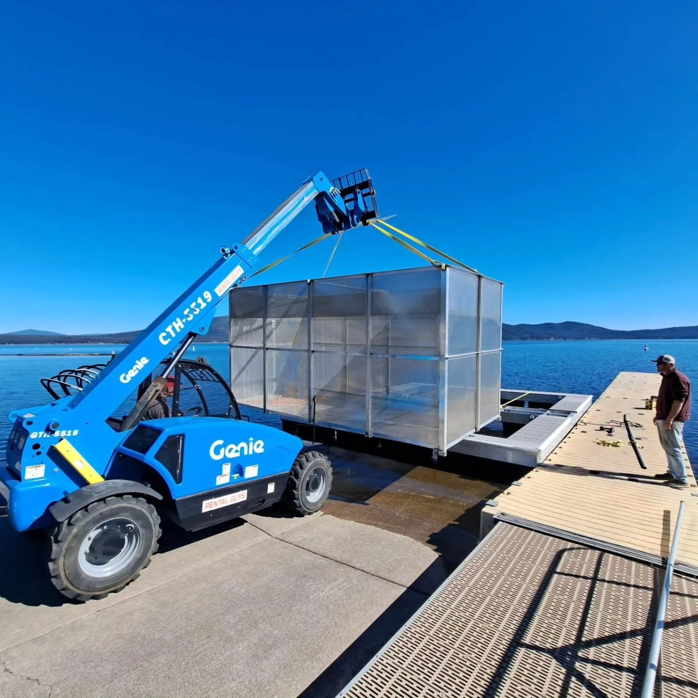 A blue Genie telescopic lift is transferring a large glass and metal structure onto a dock by the water, with a man standing nearby watching. The scene is set against a calm lake with mountains in the background on a clear, sunny day.