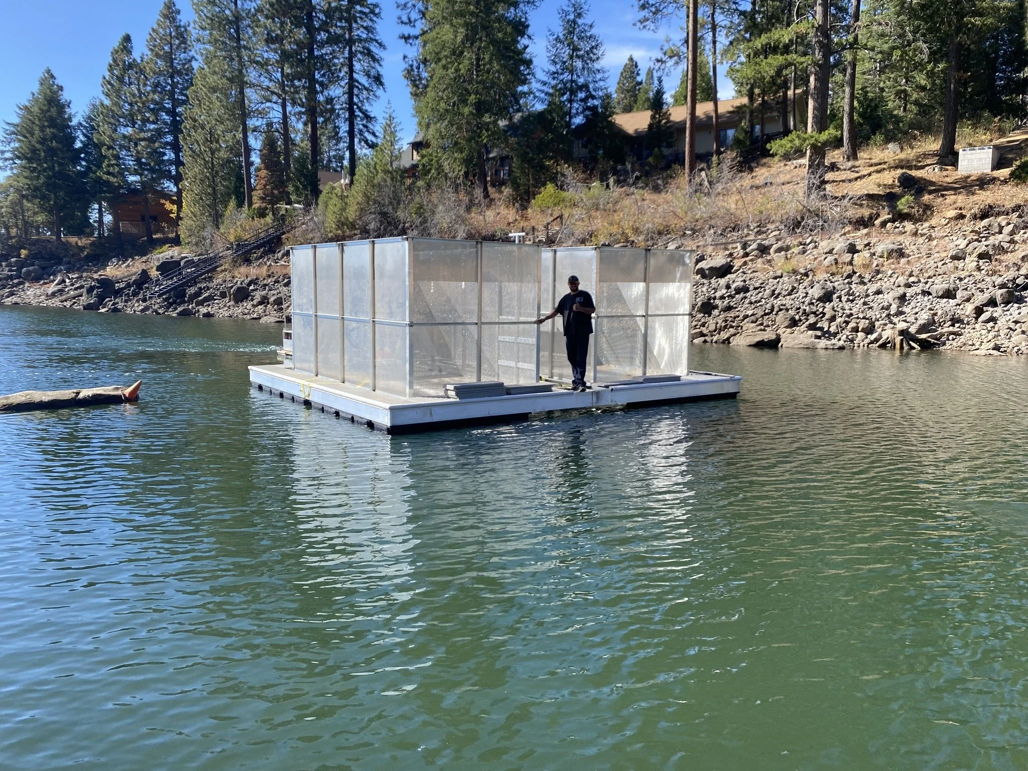 Person standing on a floating platform with clear panels on a lake, surrounded by pine trees and residential houses on a hillside.