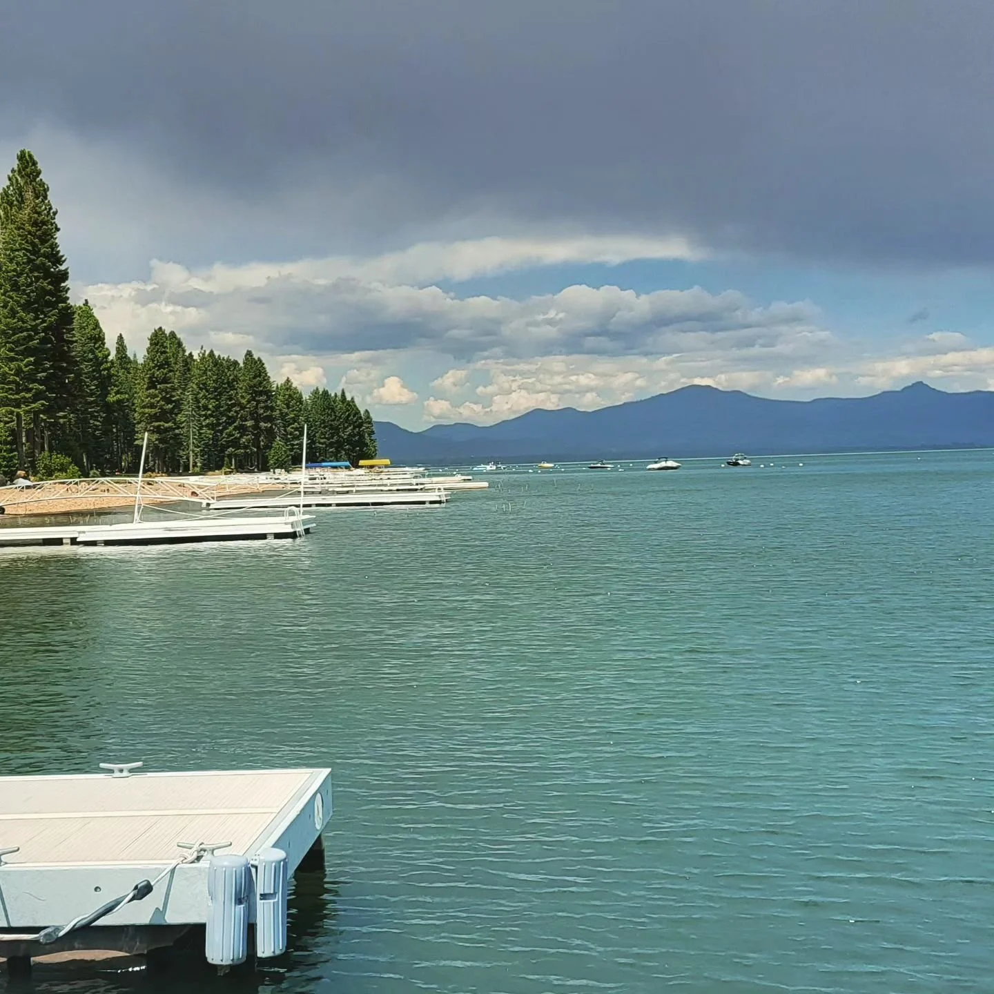 A lakeside scene with docked boats, pine trees, and mountains in the background under a partly cloudy sky.