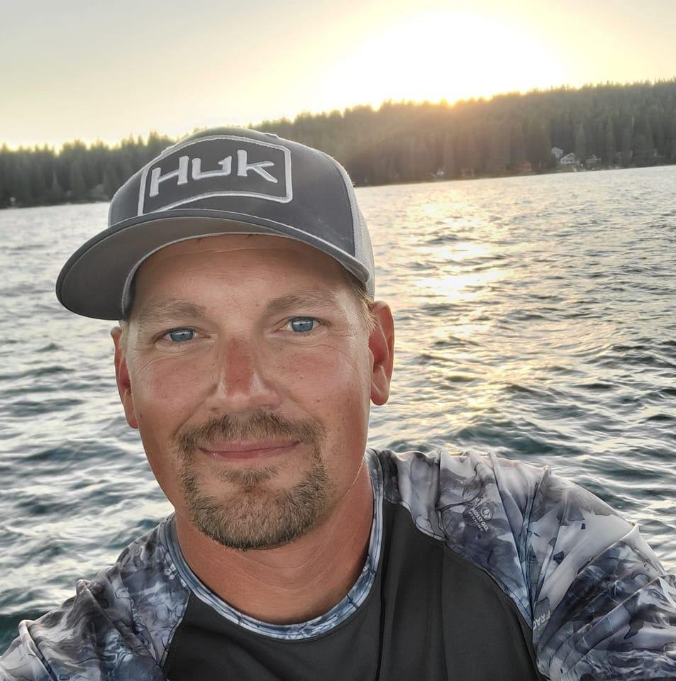 A man with a goatee, wearing a gray Huk cap and a gray patterned fishing shirt, taking a selfie on a lake during sunset with trees and a few houses in the background.