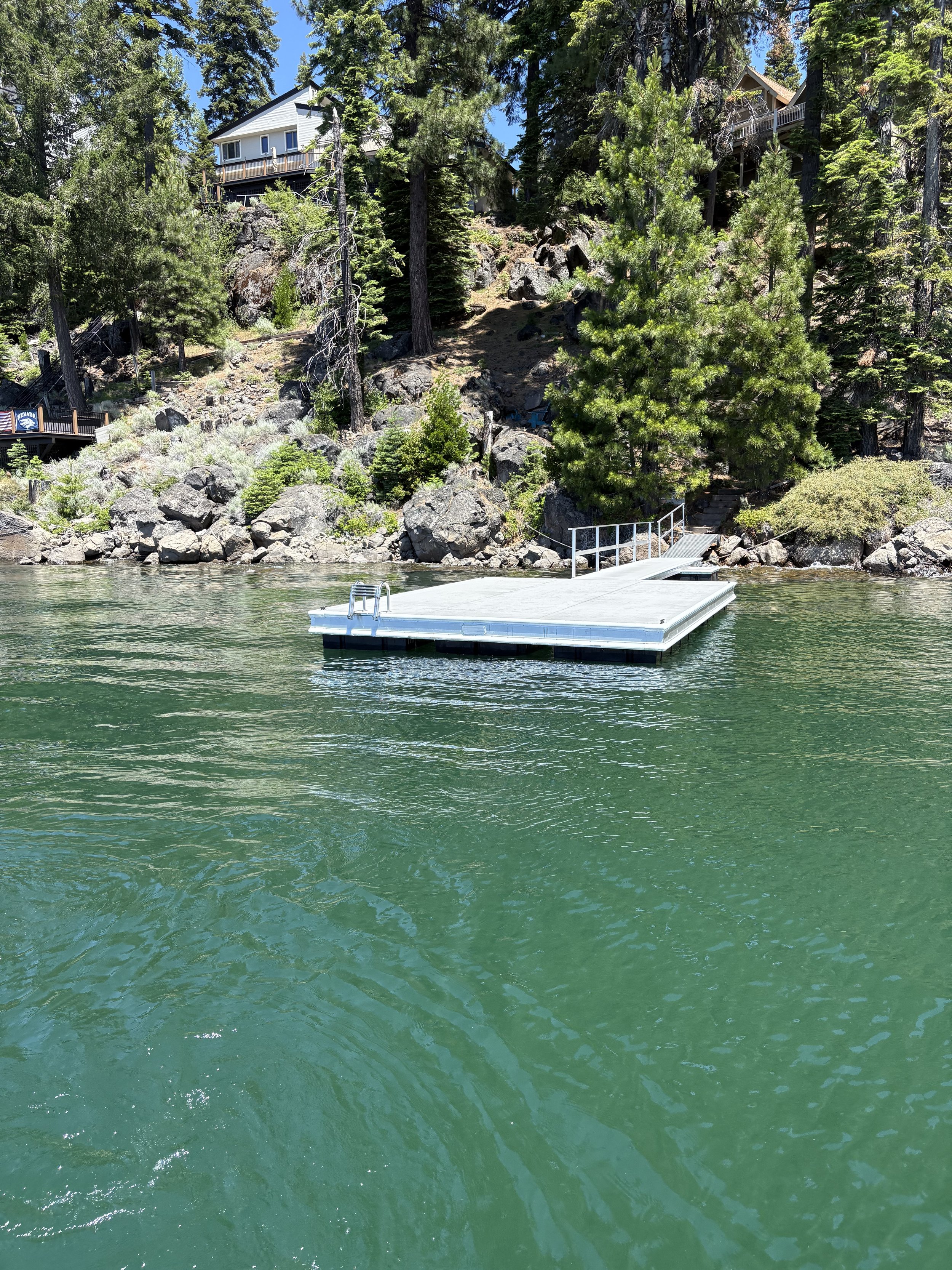 A dock floating on a body of water with trees and houses on a rocky hillside in the background.
