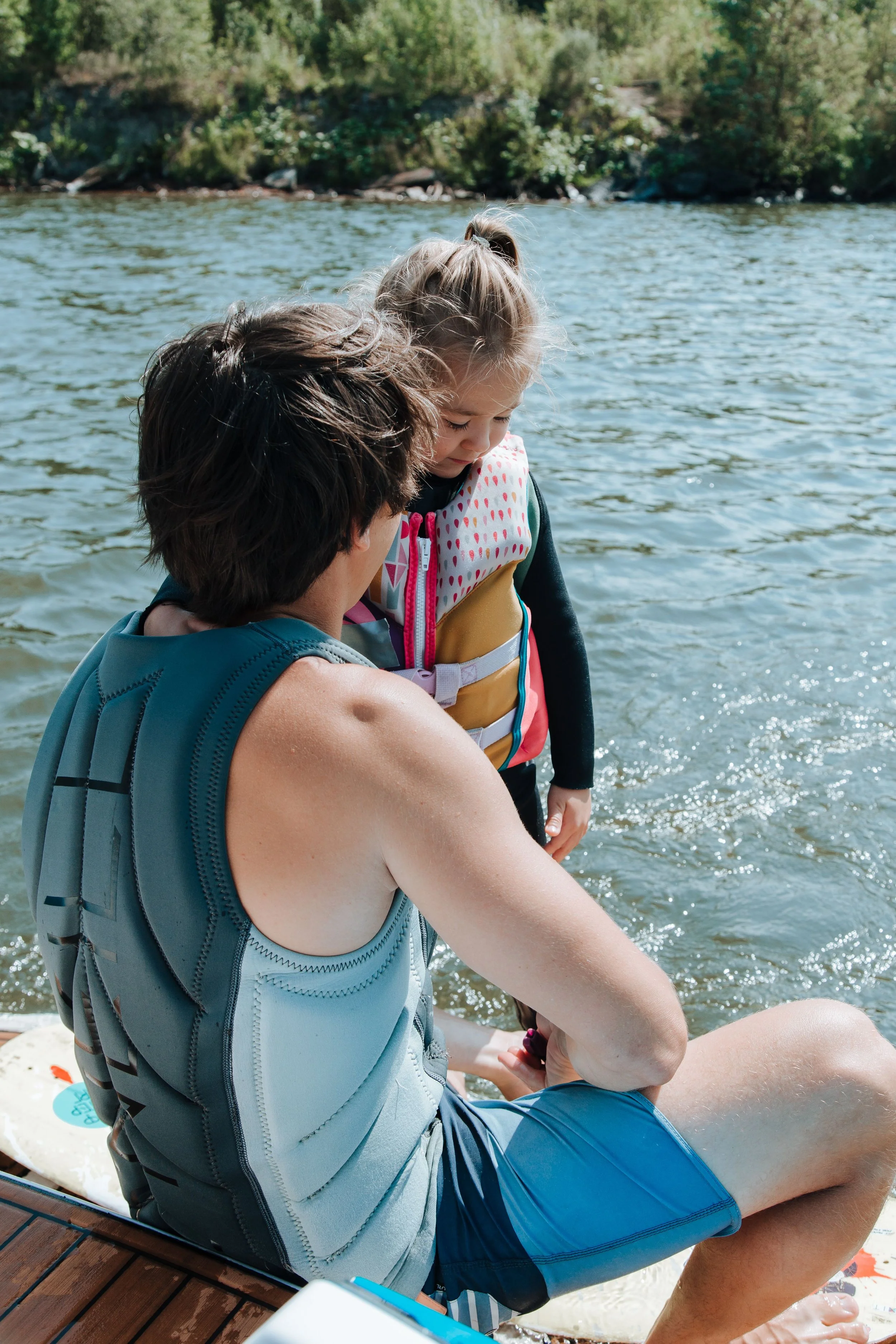A man and a girl sitting by the water, with the girl looking down and the man supporting her. The girl wears a colorful life jacket, and the man wears a sleeveless shirt and has his arm around her. They are on a wooden dock near a lake or river with trees in the background.