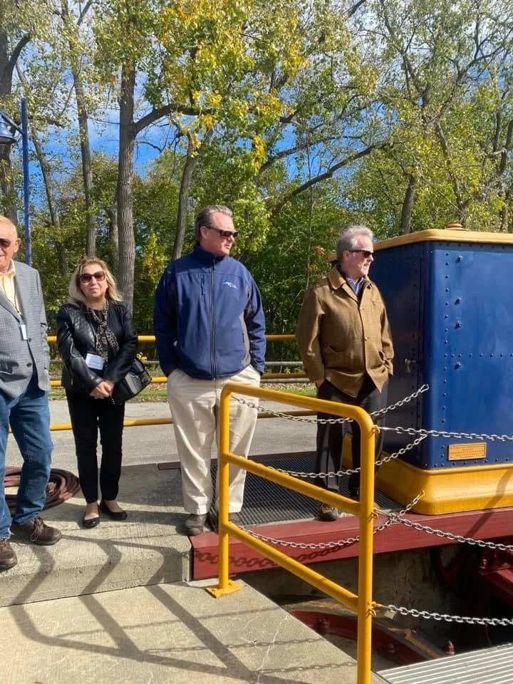 People standing in line outdoors near a blue and yellow train car. The background shows trees with fall foliage, and the sky is partly cloudy.