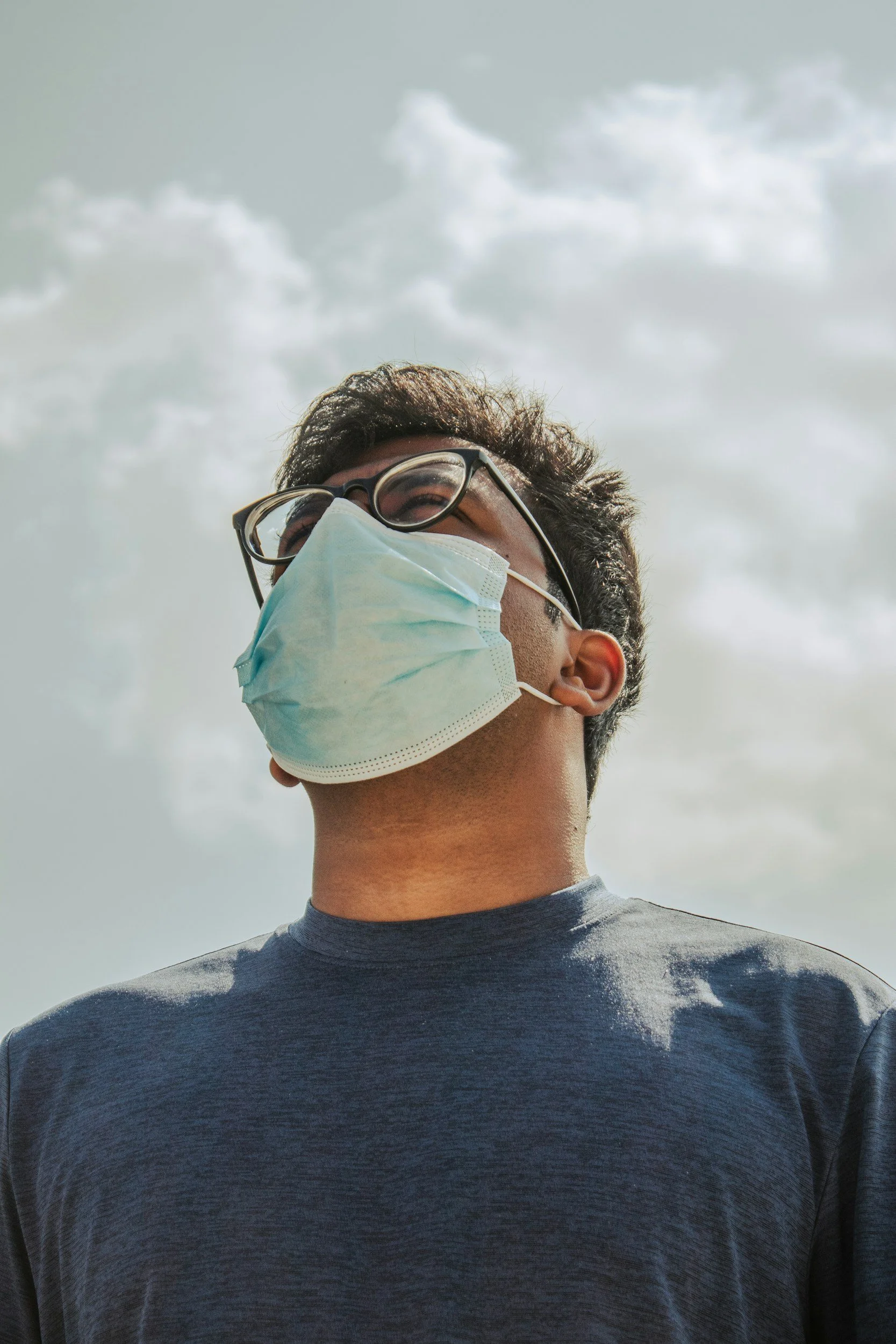 A person with glasses wearing a face mask looking upward, with a cloudy sky in the background.