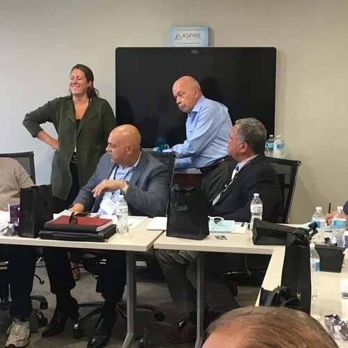 Four people in a meeting room, with tablets, water bottles, and papers on the table. Two men are seated at the table, one standing behind a large monitor, and a woman stands smiling nearby.
