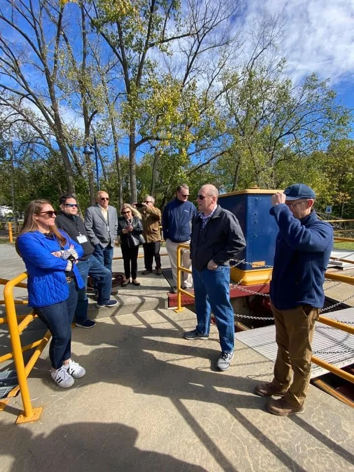 A group of people standing outdoors on a sunny day with trees in the background. They are gathered around a blue and yellow structure, possibly part of an industrial or recreational setup.