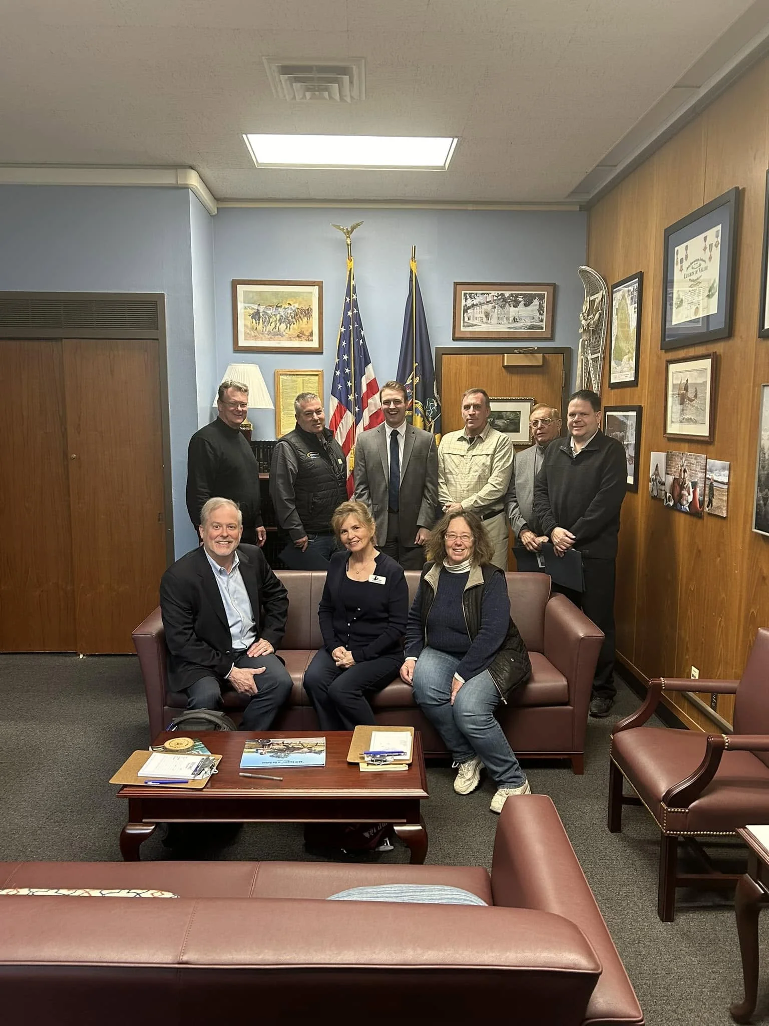 Group of people gathered in a room with wooden and blue walls, framed pictures, two flags, and a reception desk.