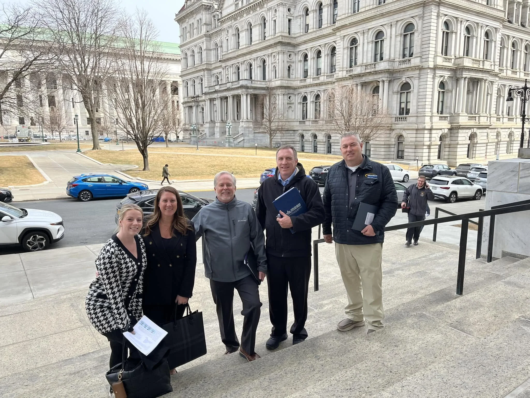 A group of people smiling and standing outdoors on a city sidewalk in front of a large historic building with detailed architecture. The group includes men and women holding papers and folders, with parked cars and leafless trees in the background.