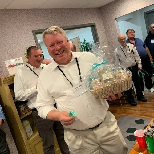 Man smiling and holding a gift basket with cookies at a gathering of people indoors.