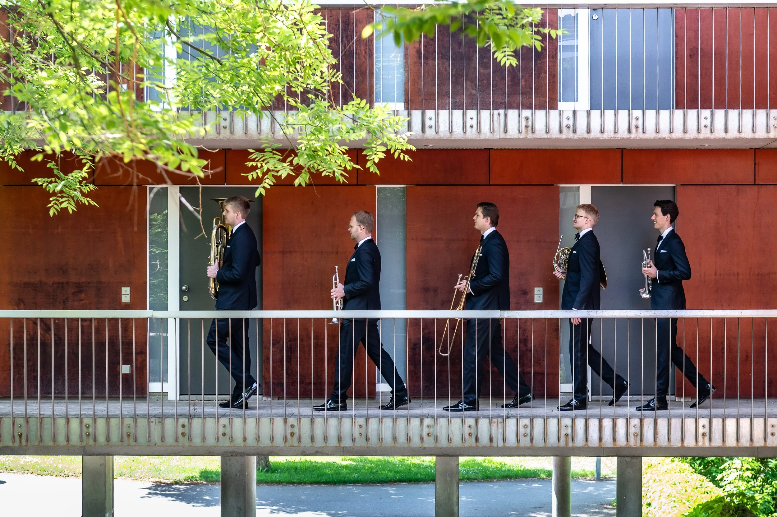 Five musicians in formal black suits playing brass instruments while walking along a sidewalk with a modern building and green foliage in the background.