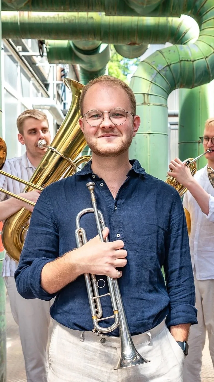 A man in a blue shirt holding a trumpet, standing in front of two men playing brass instruments, in an industrial or garden setting.