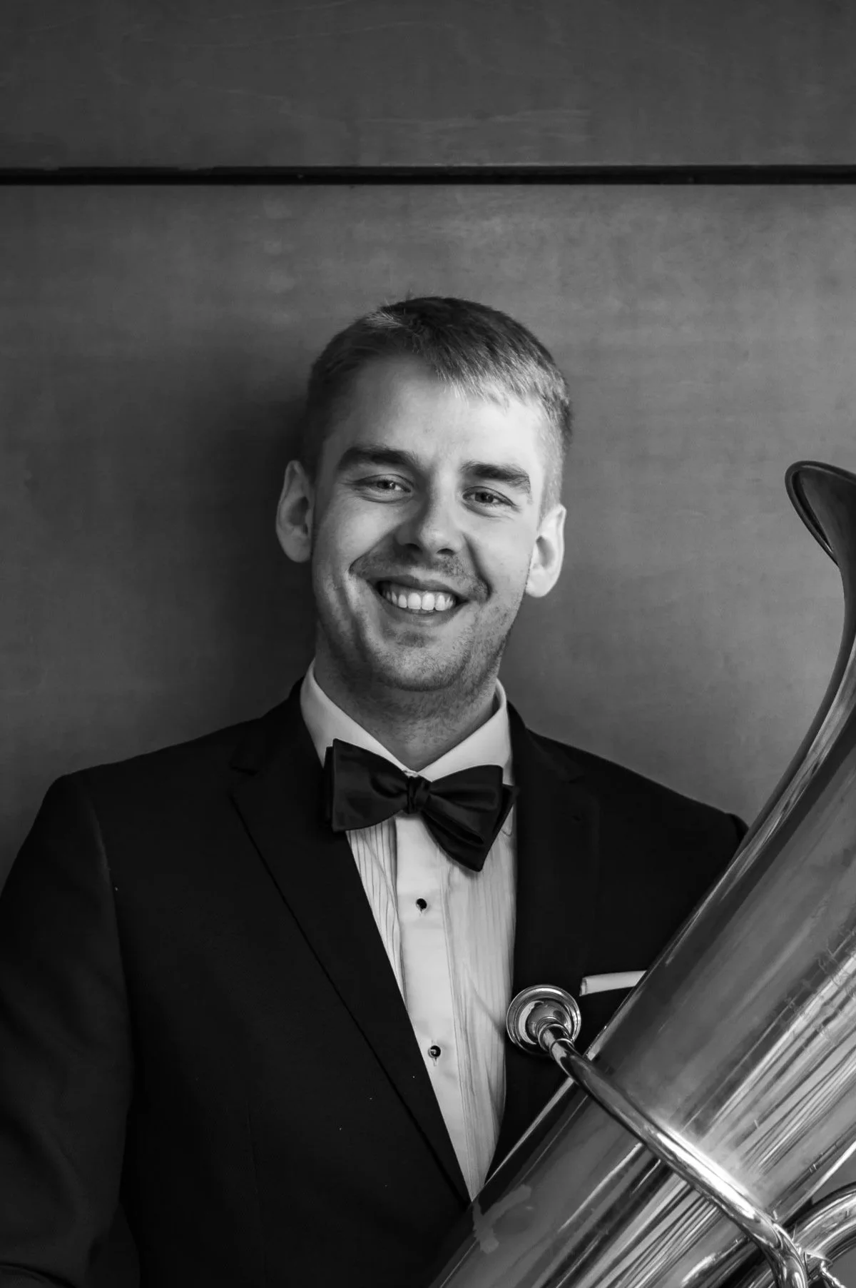 A smiling young man in a tuxedo and bow tie holding a musical instrument, possibly a tuba, in front of a plain wall.