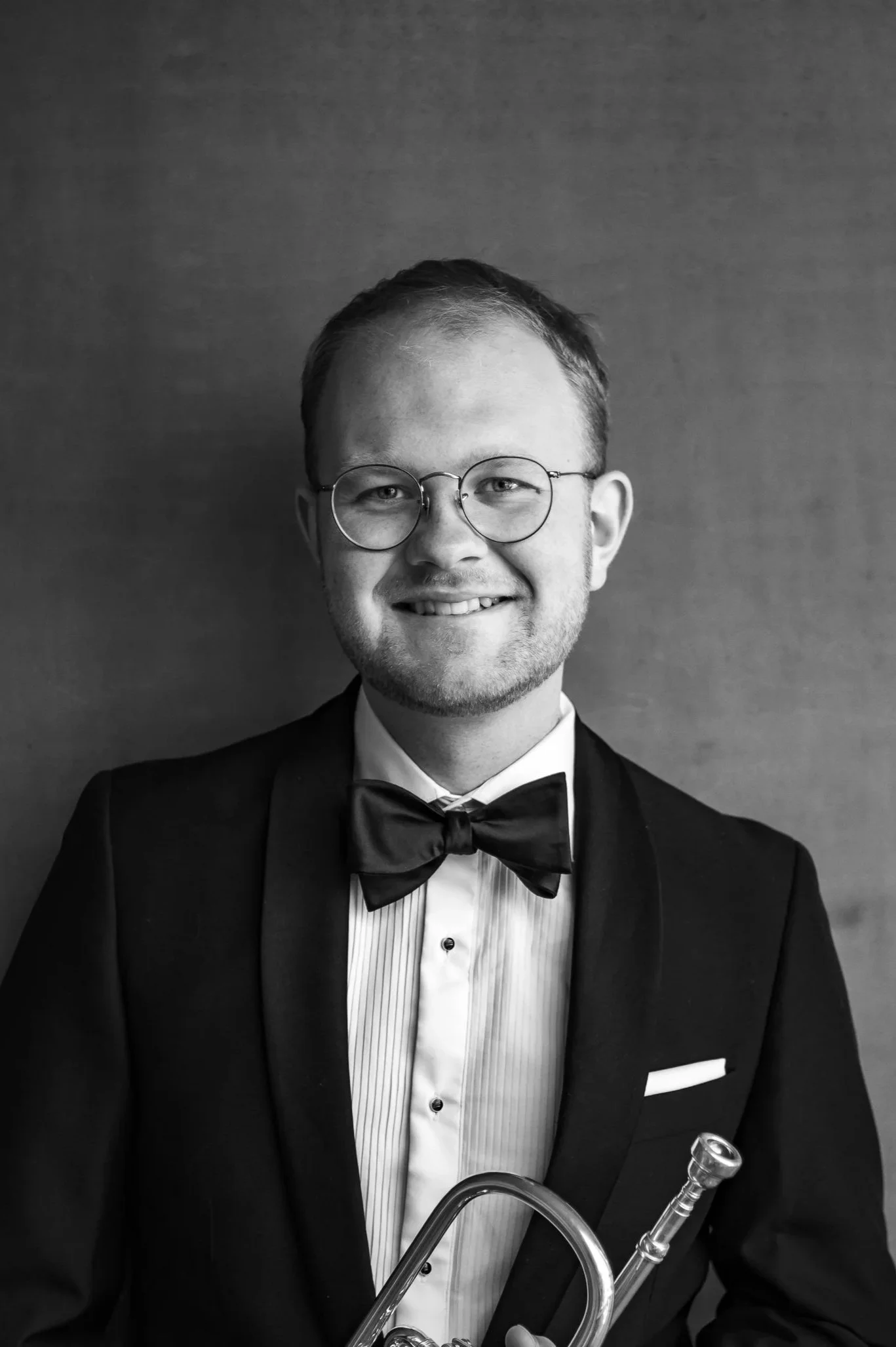 Black and white photo of a man dressed in a tuxedo, wearing glasses, holding a trumpet, standing against a plain background.