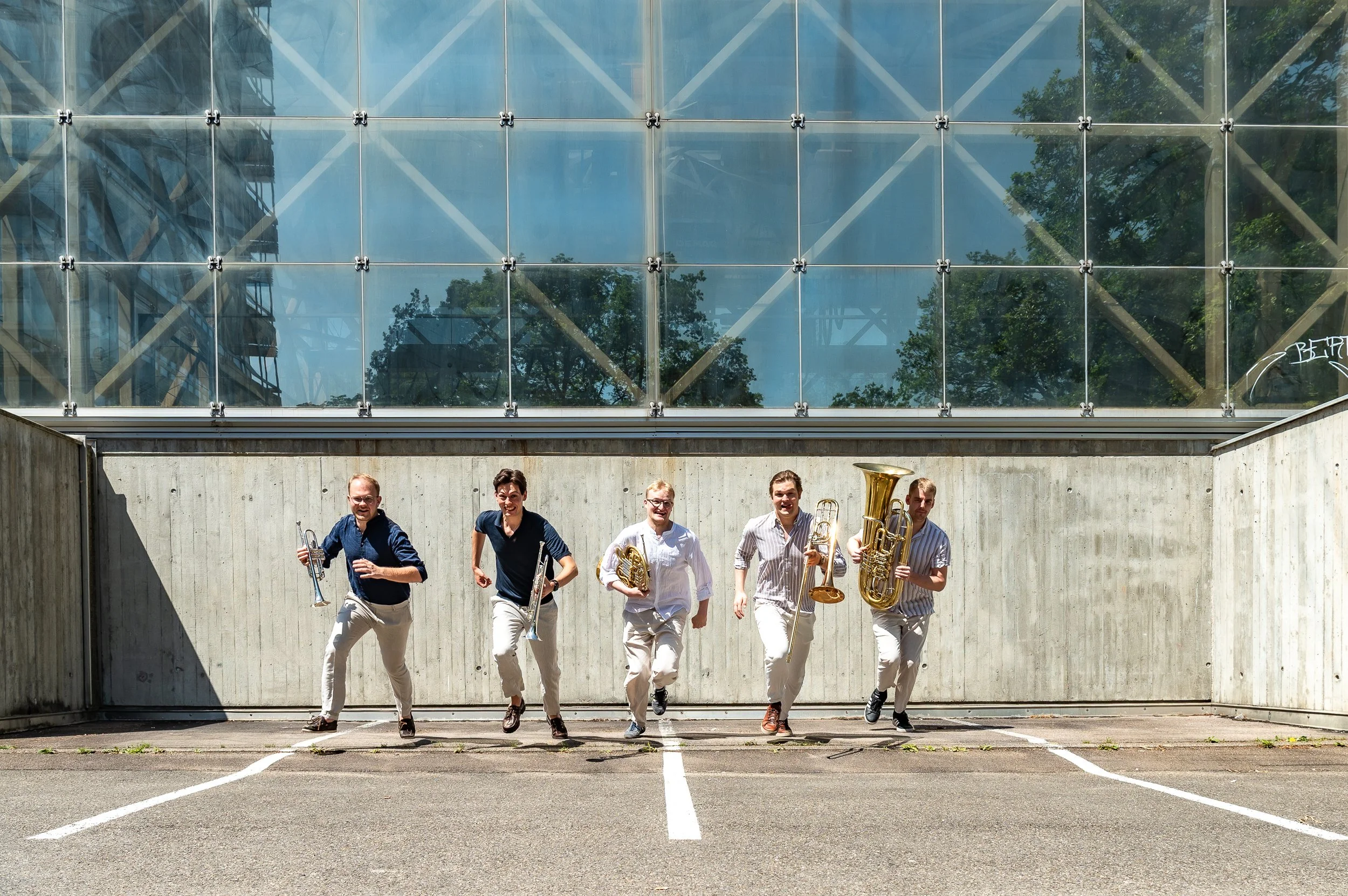 Group of five men walking and running outdoors in front of a modern building with glass panels, some holding musical instruments like a trumpet and a tuba, on a parking lot with marked parking spaces.