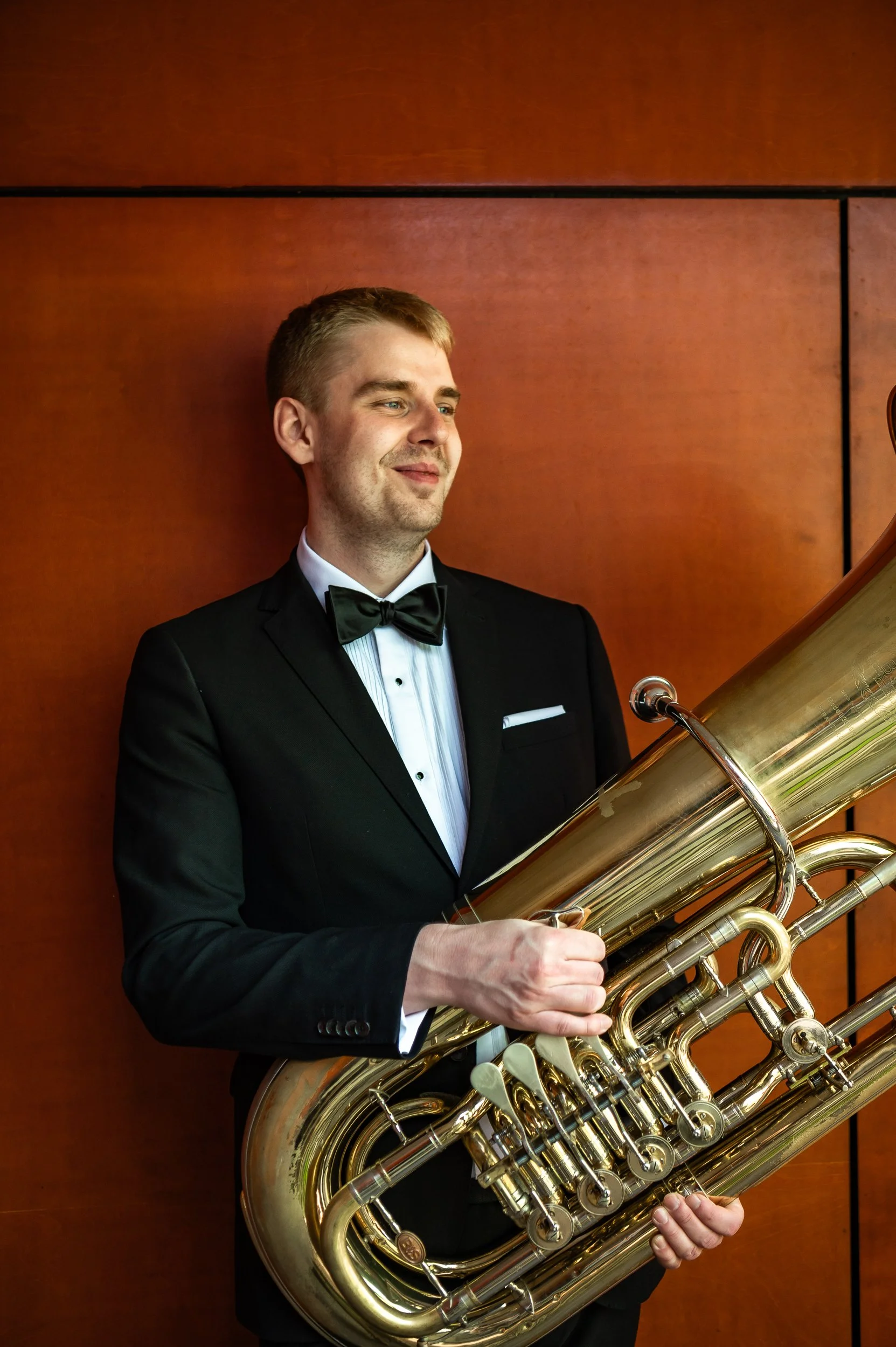 A young man in a tuxedo playing a large brass tuba, standing against a wooden wall.