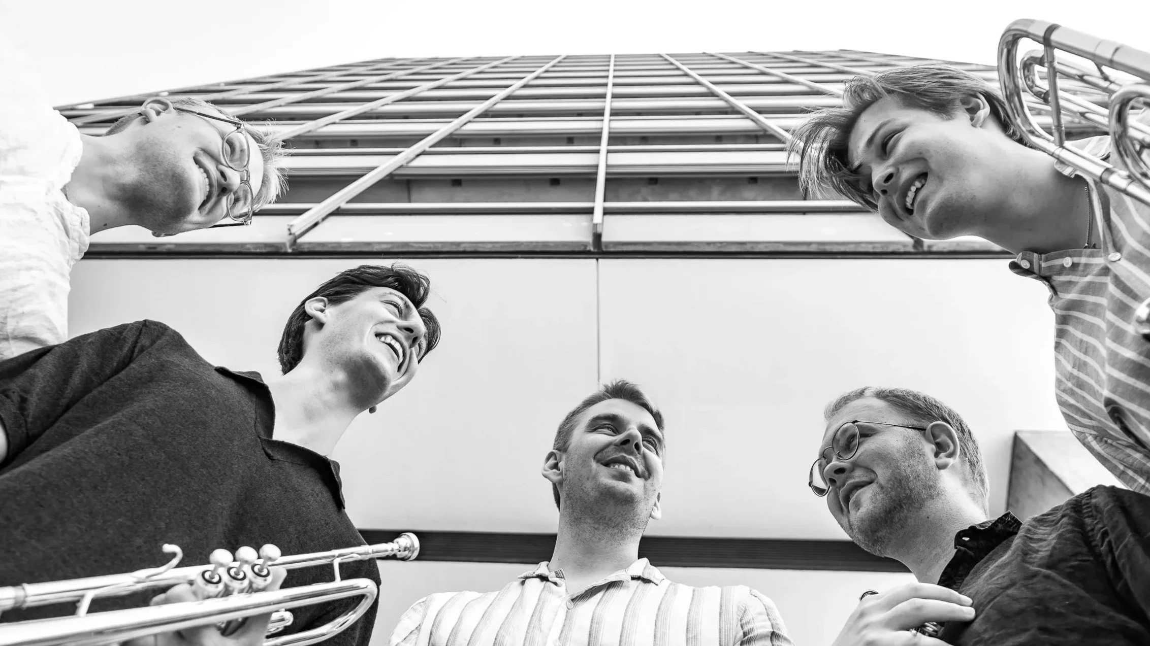 Six young adults smiling and talking in a circle outdoors, with a tall building in the background.