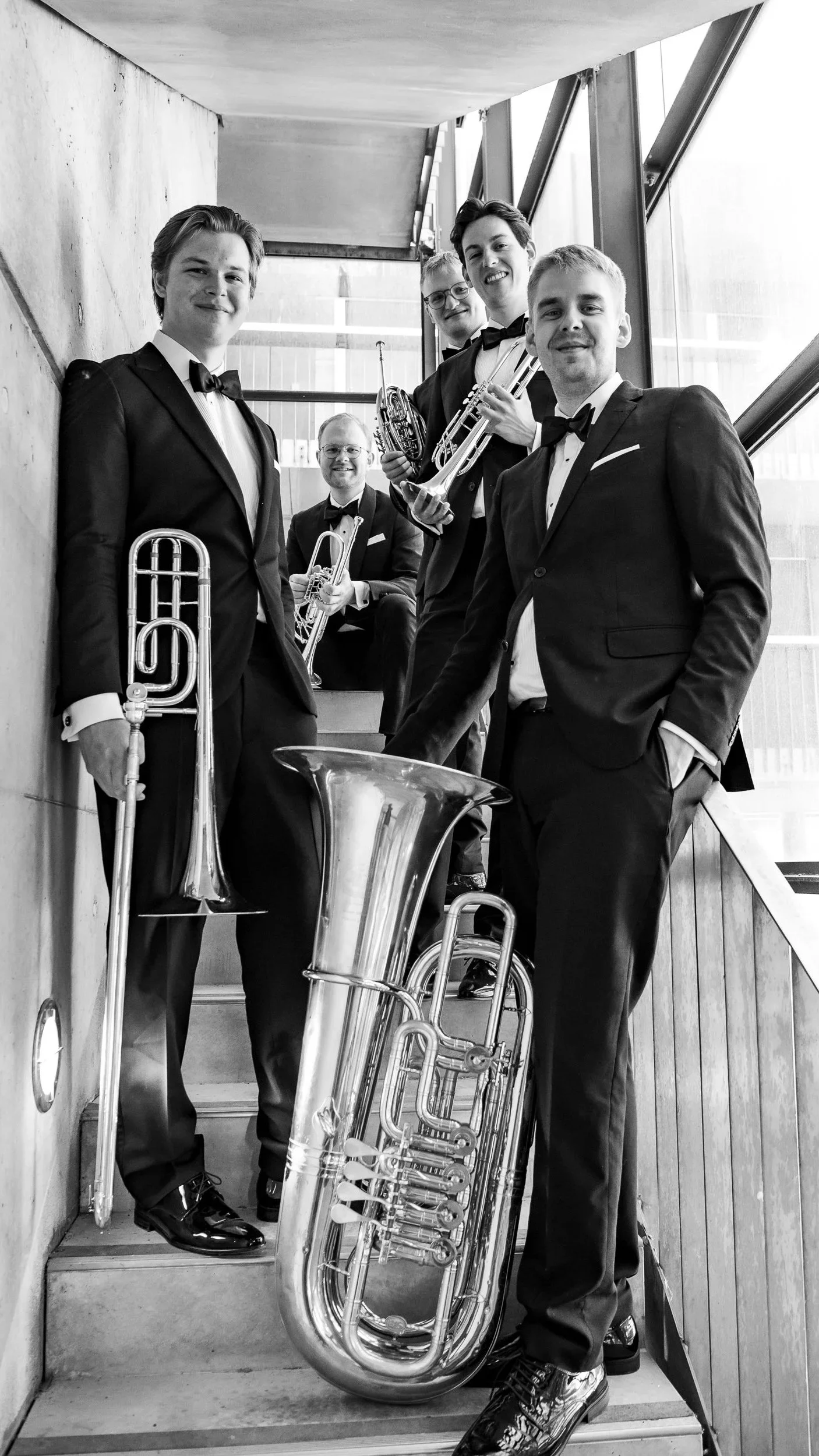 Black and white photo of five young men in tuxedos with bow ties, holding brass instruments, standing on a staircase near a concrete wall and large window.