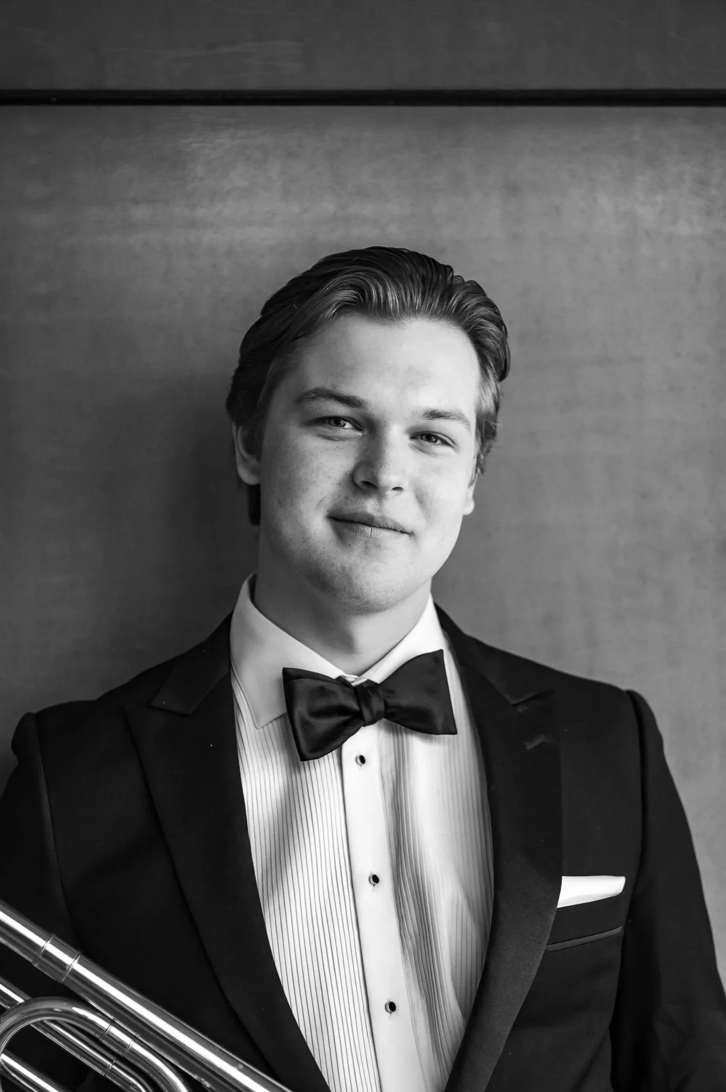 A young man in a tuxedo with a bow tie holding a flute, standing against a plain wall.