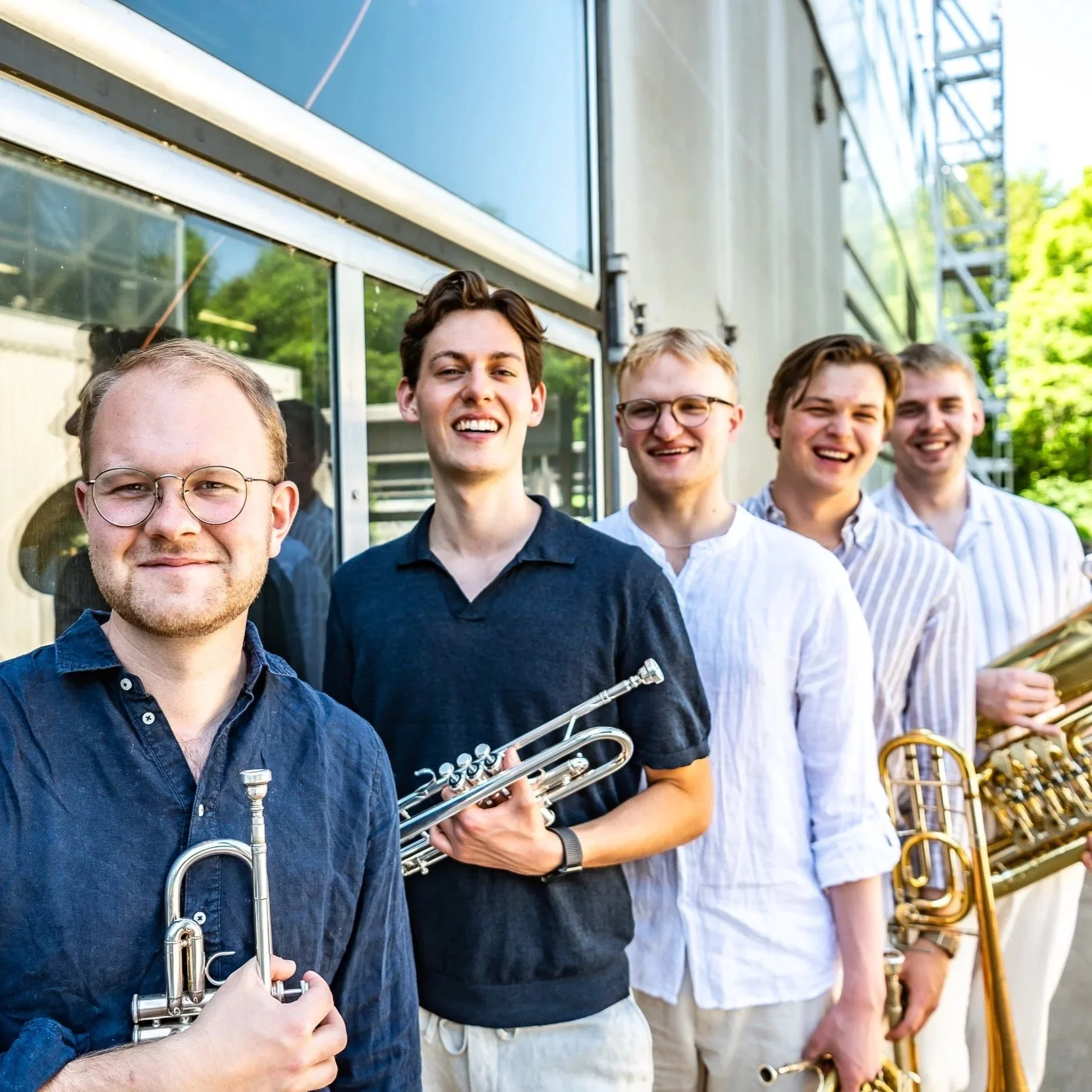 A group of five young men standing outdoors next to a building with glass windows, holding musical instruments, with trees and a metal staircase in the background.
