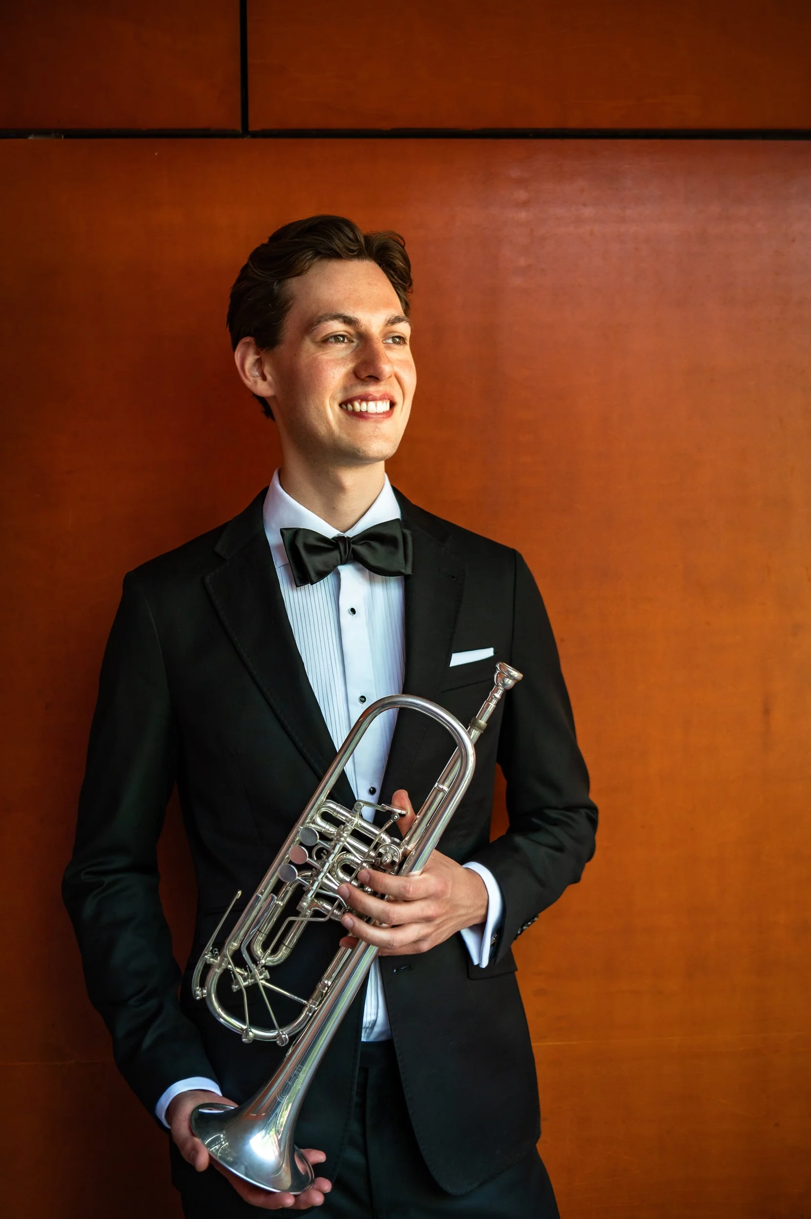 A young man in a black tuxedo, white shirt, and black bow tie holding a silver trumpet, standing in front of a wooden wall.
