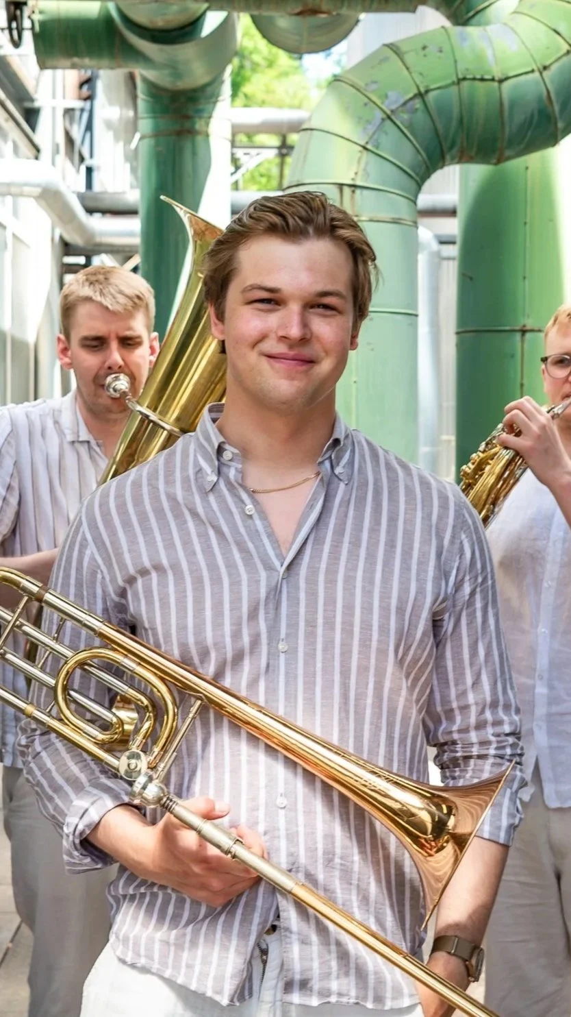 Young man smiling and holding a gold saxophone, with two other musicians playing brass instruments in the background, outdoors with green pipes.