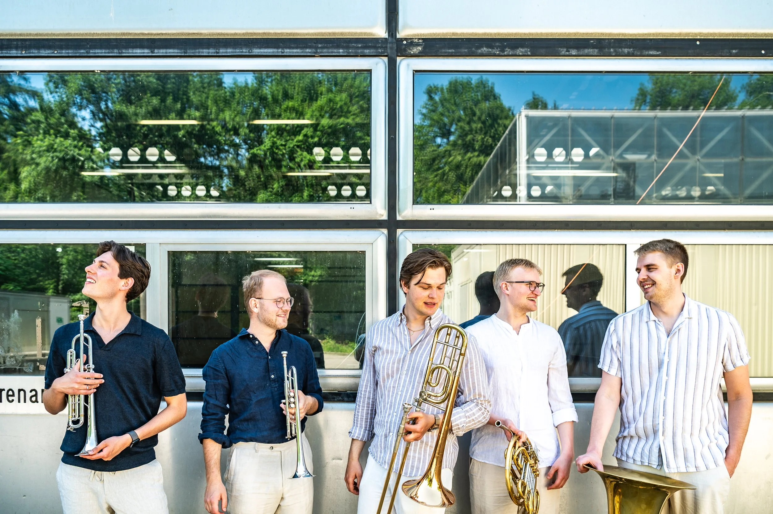 Group of five young men standing outside in front of a building with reflective windows, holding musical instruments including trumpets and a trombone, smiling and engaging in conversation.