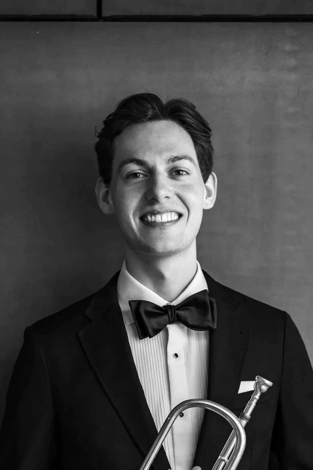 A young man in a tuxedo with a bow tie, smiling and holding a trumpet.