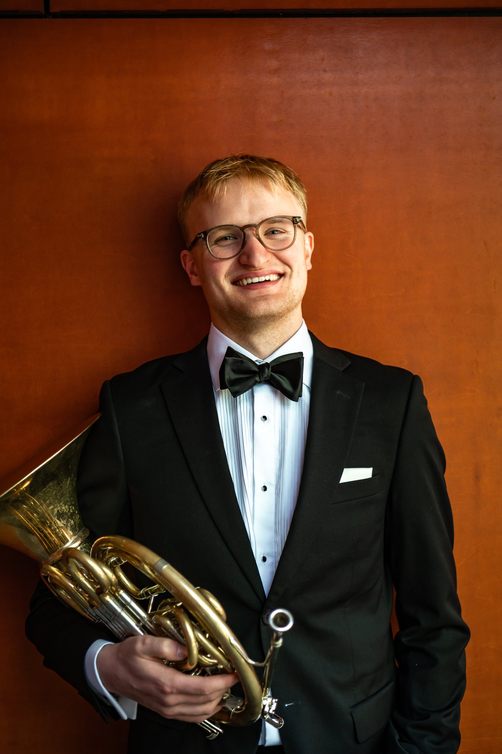 A young man in a tuxedo holding a brass musical instrument, smiling and standing against a wooden wall.