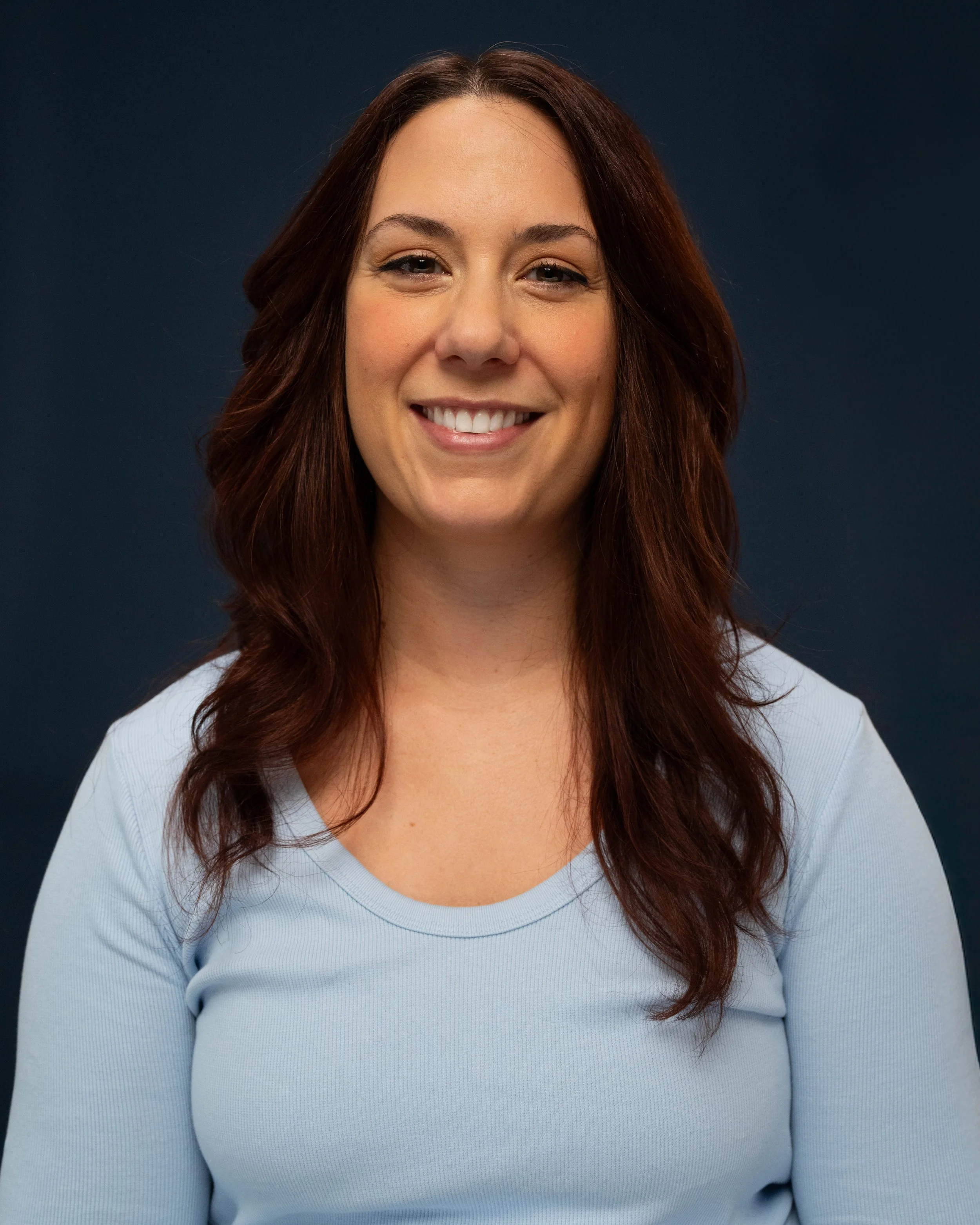 A smiling massage therapist with long, wavy brown hair wearing a light blue long-sleeve top, standing against a dark background.