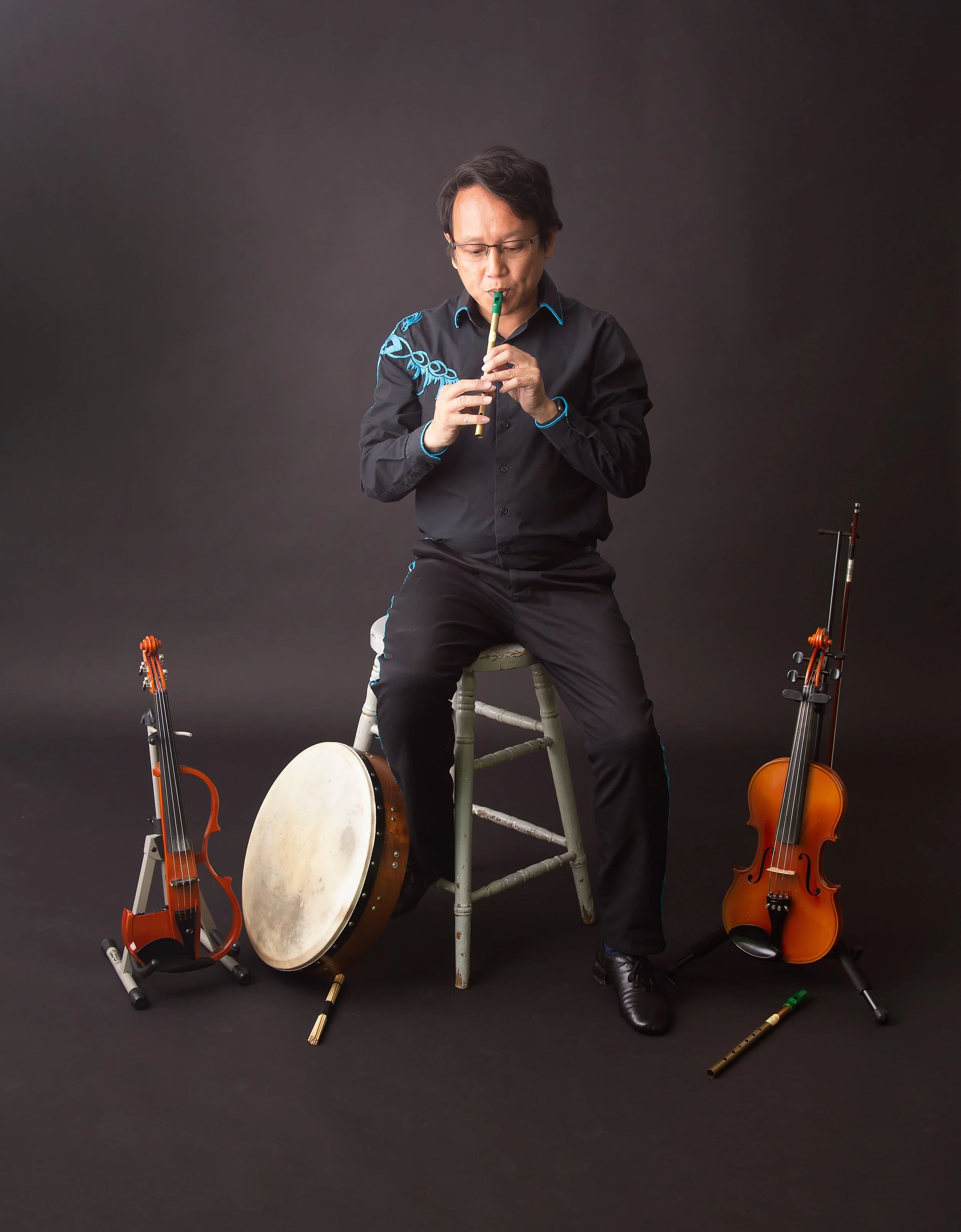 Marcel sitting on a stool playing the whistle, surrounded by an electric fiddle, whistle, bodhrán, and acoustic violin on display on the ground.