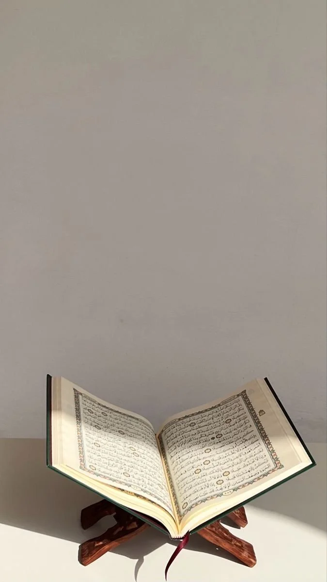 An open book with Arabic script resting on a wooden stand against a plain background.