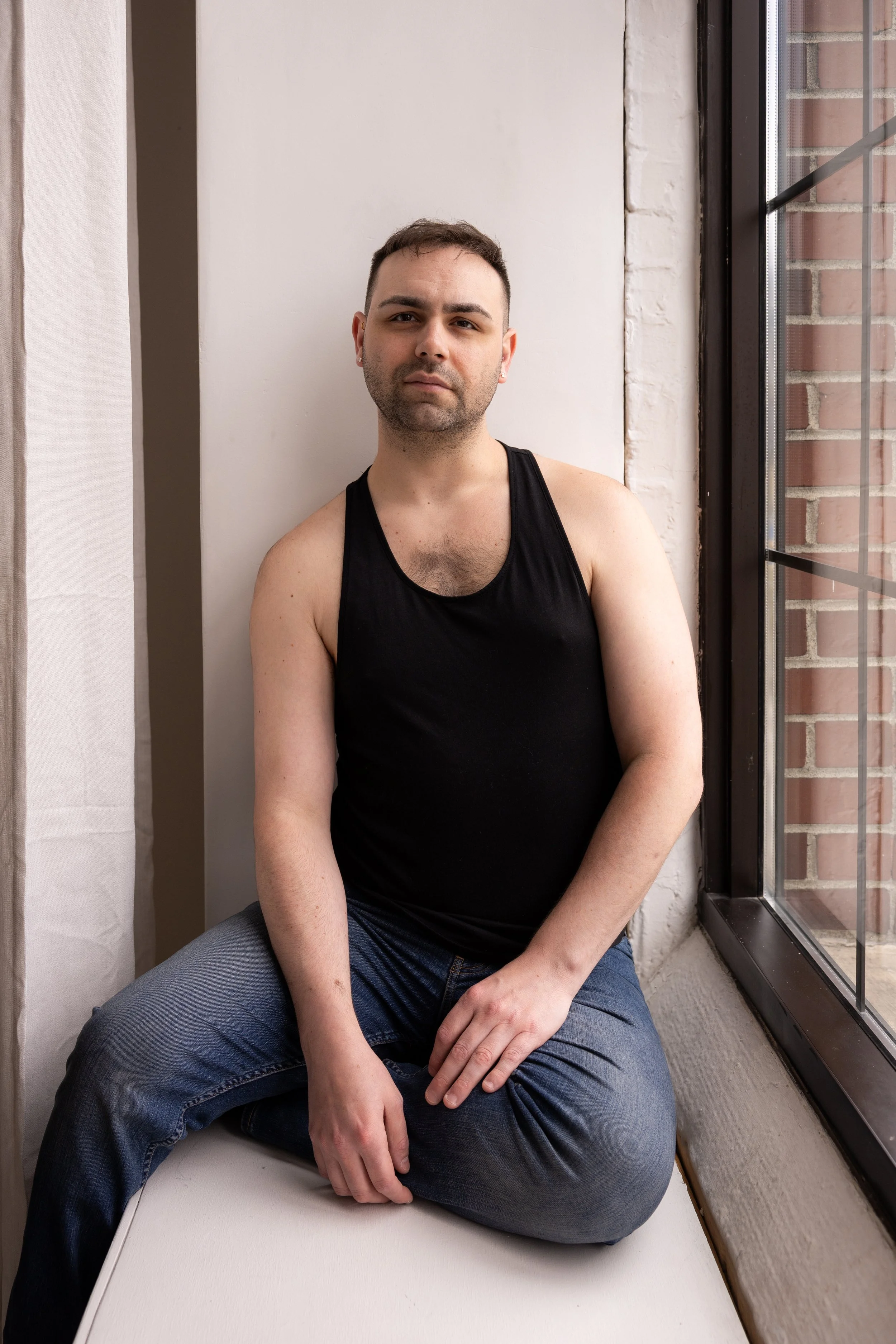 A young man with short dark hair, wearing a black sleeveless shirt and jeans, sitting on a windowsill next to a large window with brick exterior visible outside, against a white wall, looking directly at the camera with a neutral expression.
