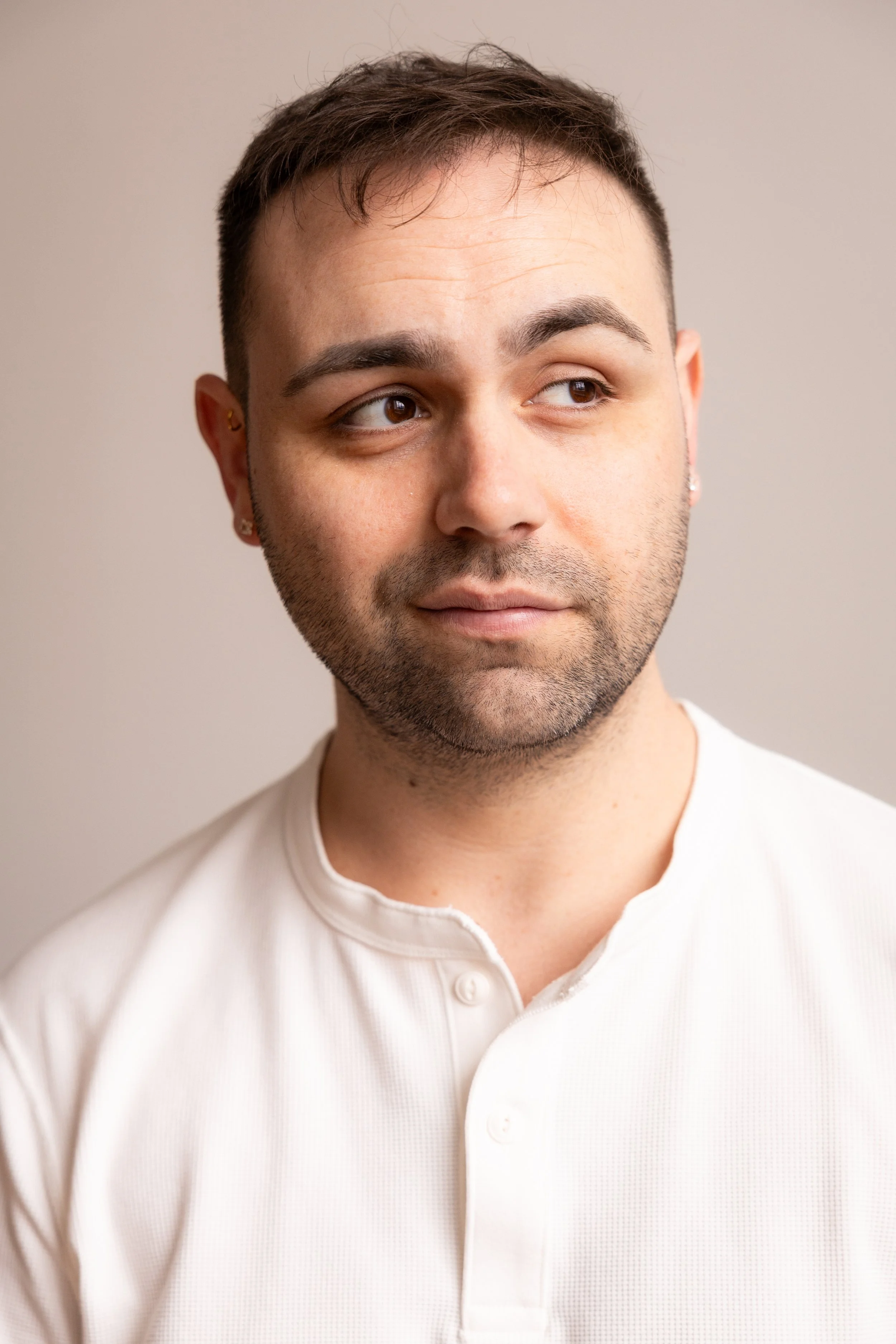 Close-up of a young man with short brown hair and light stubble, wearing a white collared shirt, looking to the side with a neutral expression against a plain background.