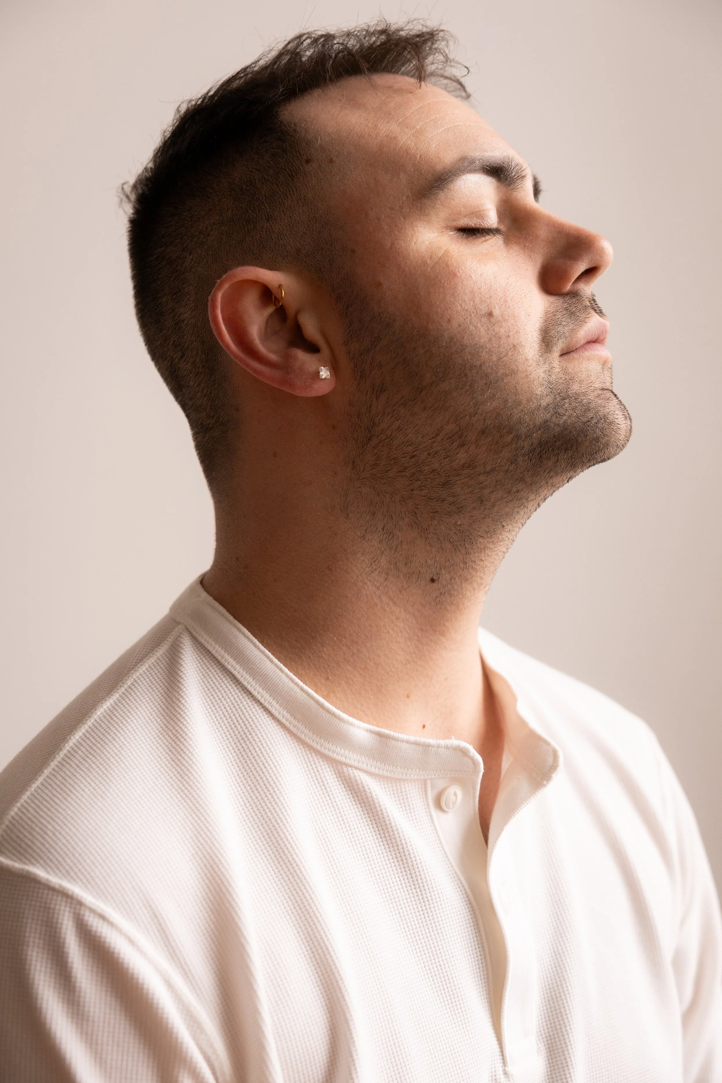 A side profile of a man with closed eyes, short brown hair, a diamond earring, wearing a white collared shirt, standing against a plain neutral background.