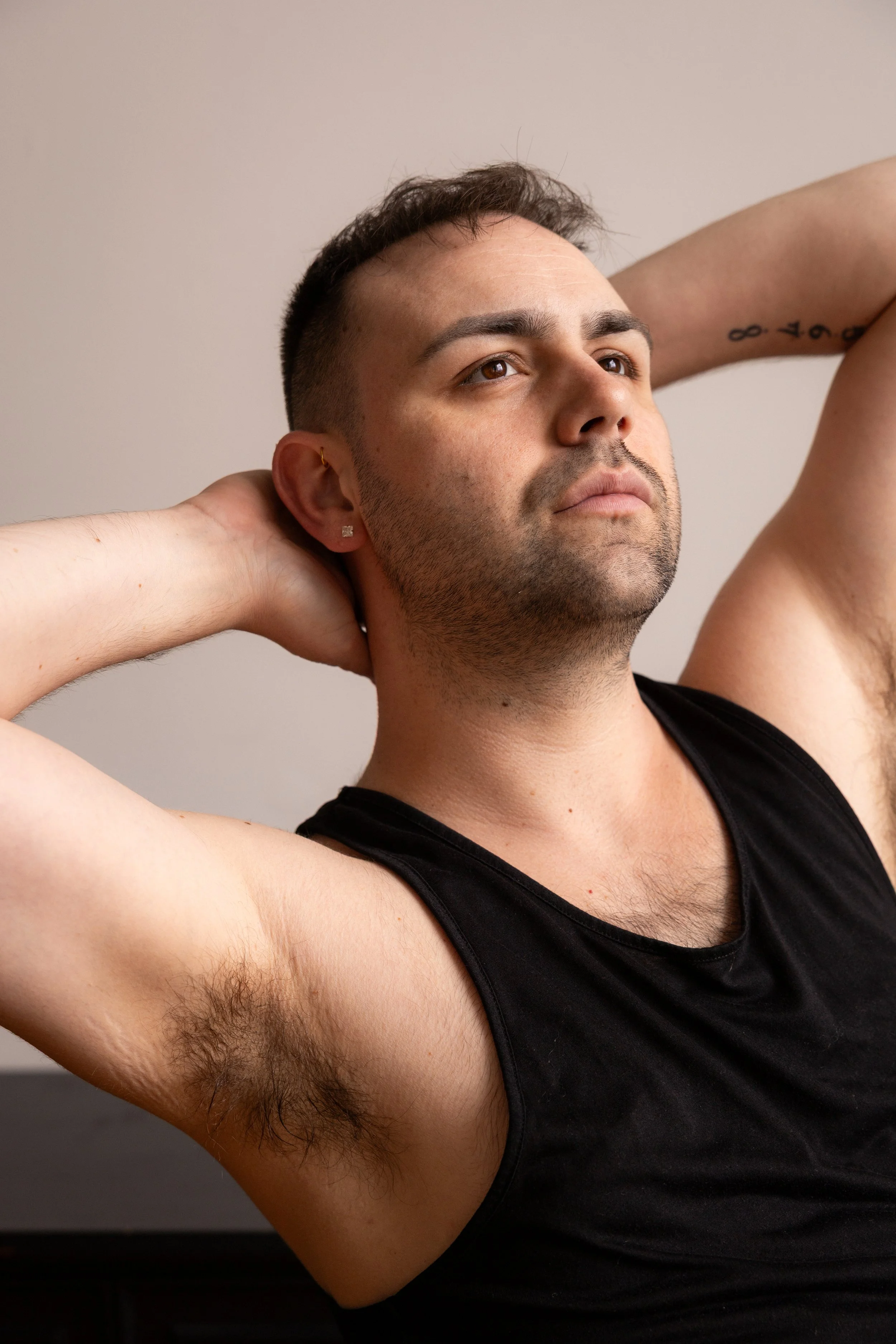 A young man with short dark hair, light tan skin, and brown eyes, wearing a black tank top, has his hands behind his head and looks thoughtfully to the side against a plain light-colored background.