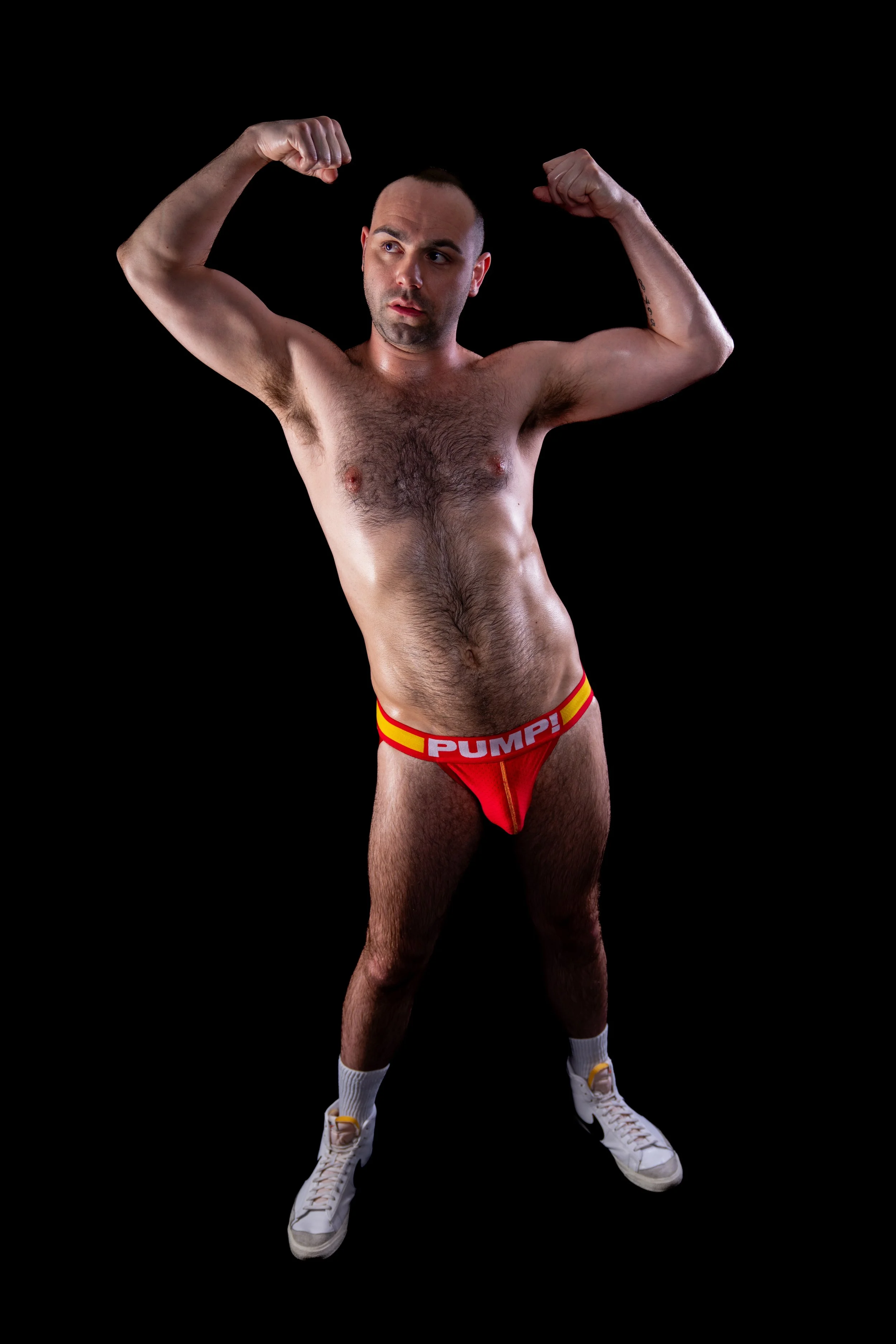 A man with a muscular build poses with both arms flexed, wearing red and yellow underwear with the word "PUMP!" on the waistband, white sneakers, and white socks, against a black background.