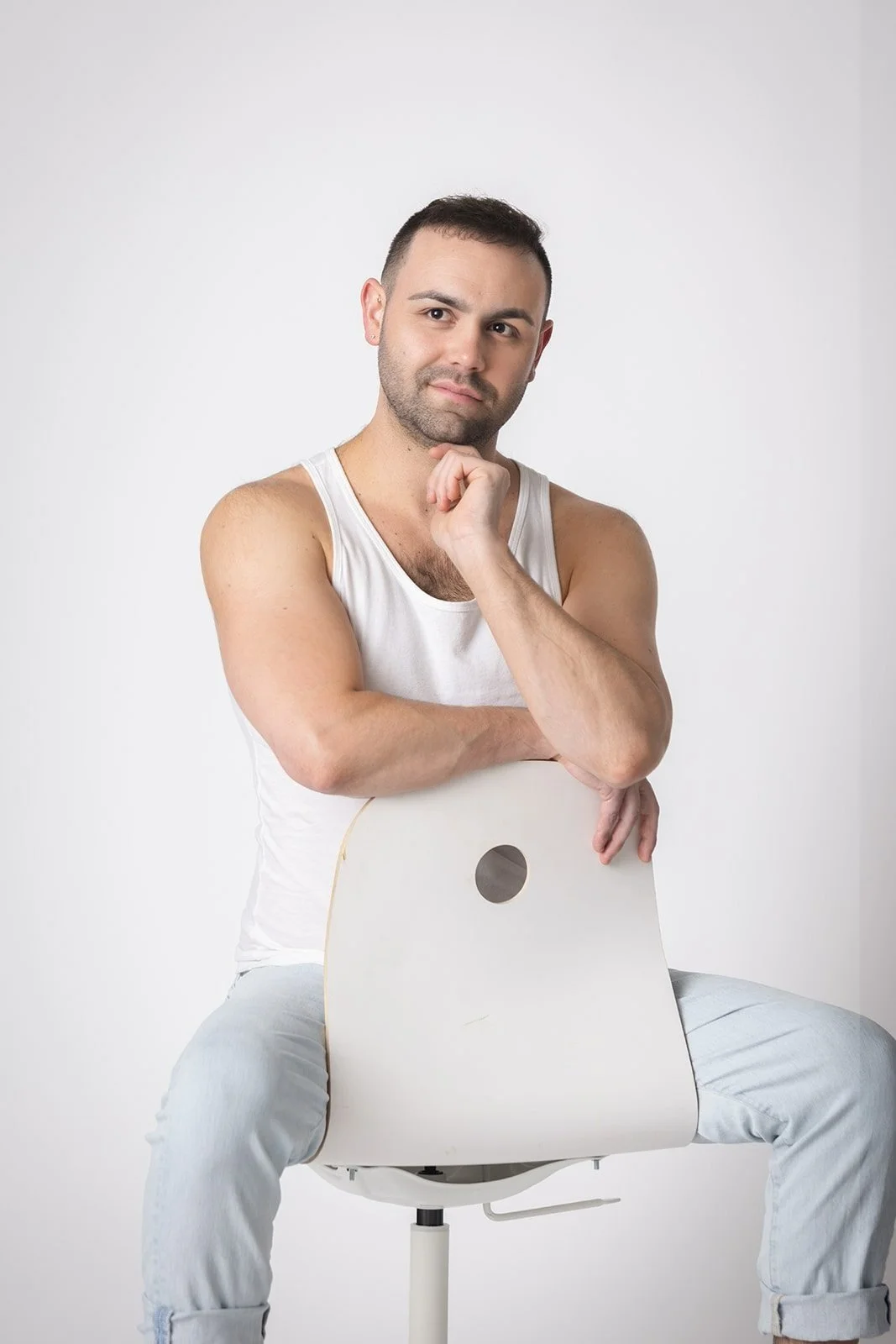 A young man sitting on a white chair, resting his chin on his hand, wearing a white tank top and light-colored jeans against a plain white background.