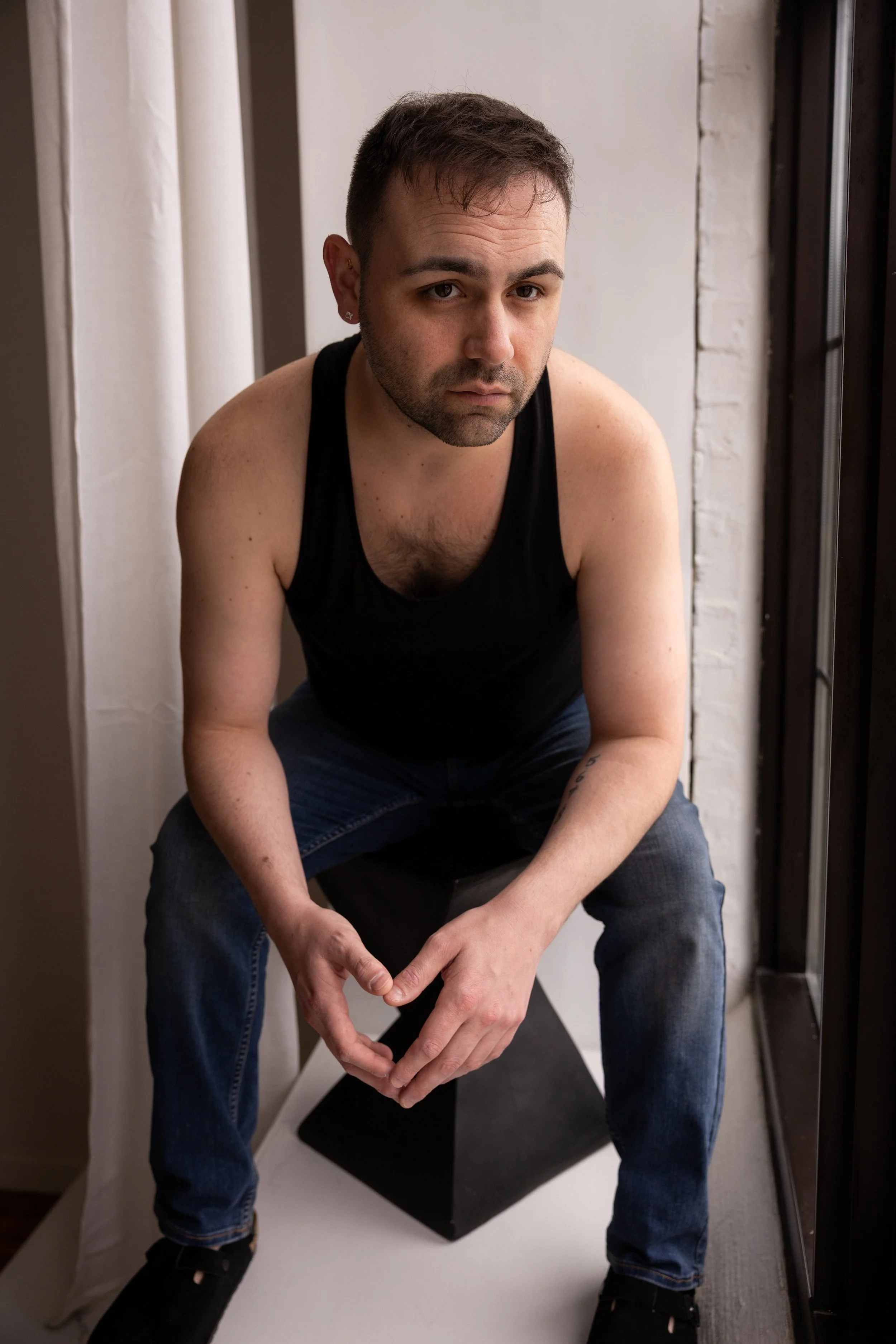 A man with short dark hair and a beard, wearing a black tank top and jeans, sitting on a black stool with a white background and a window to his right.