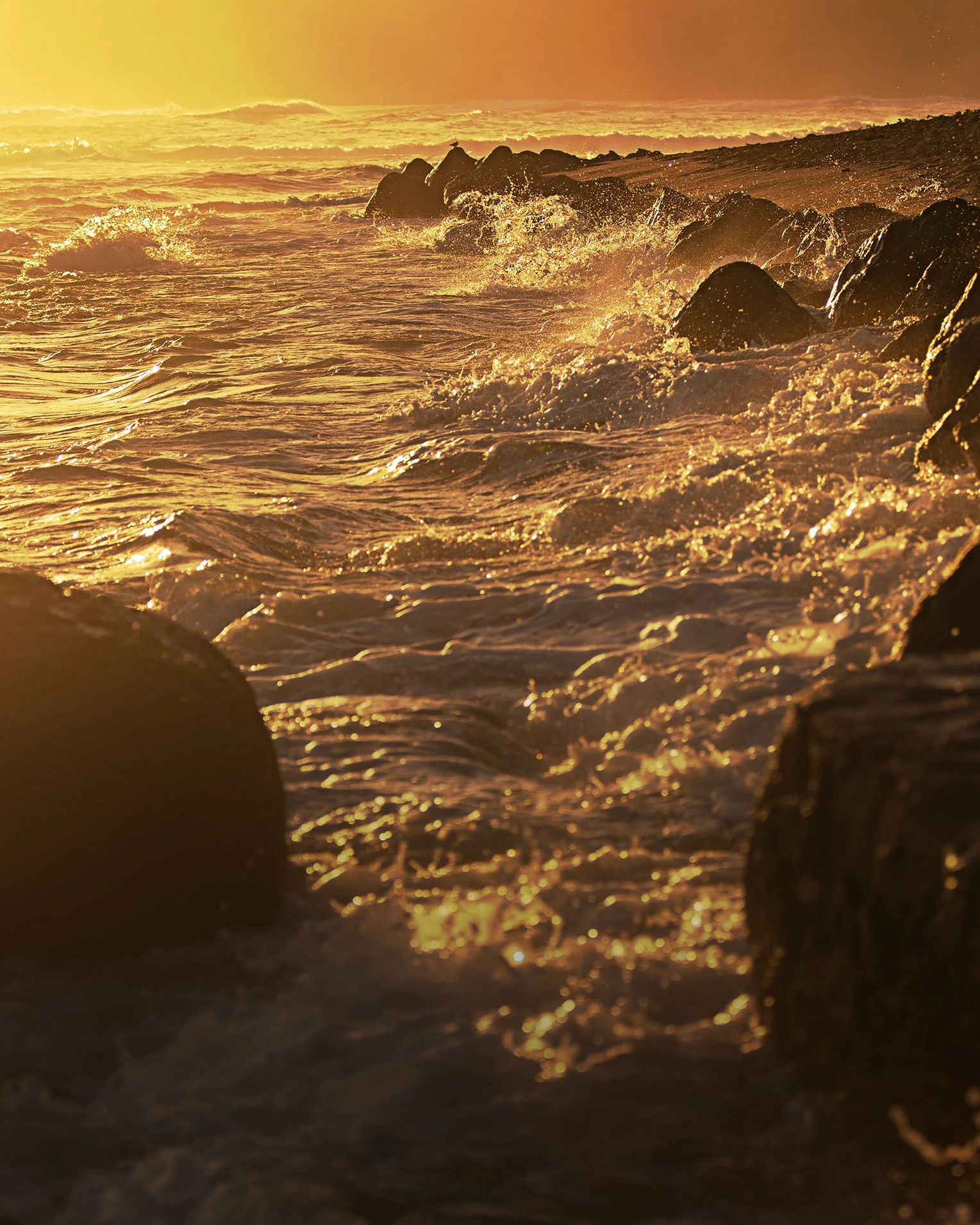 A golden sunset over the ocean with waves crashing against rocks on the shore.